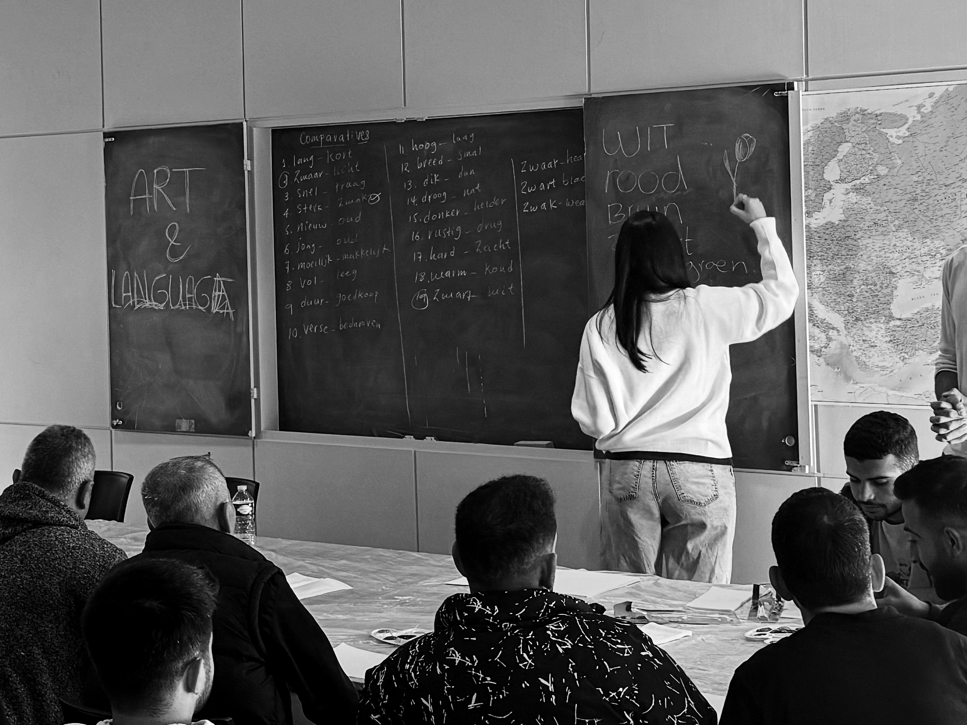 A woman teaching a class at a blackboard with students seated around a table, writing on the board and taking notes.