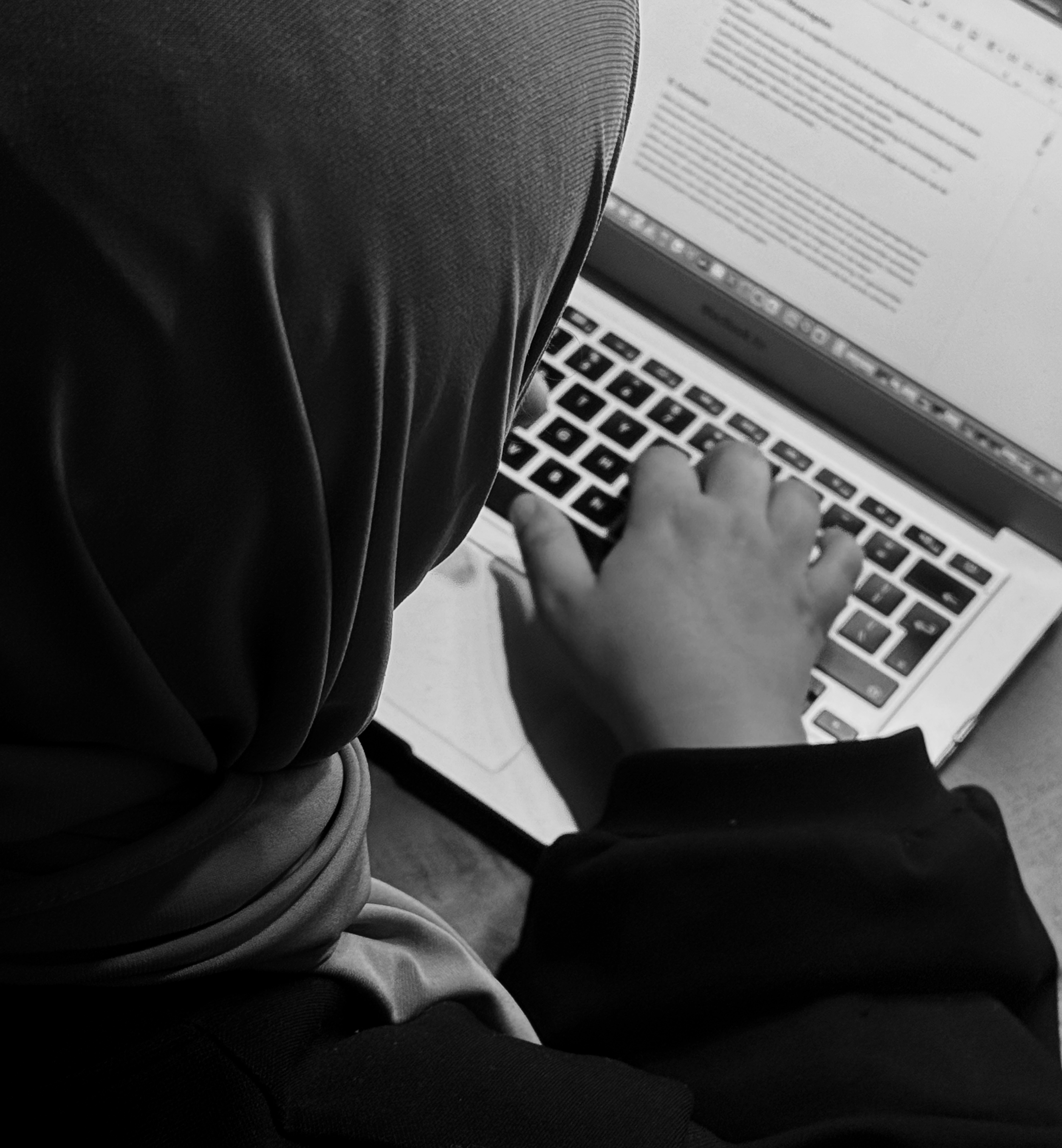 Person using a laptop, typing on the keyboard with a document open on the screen, shot from above in black and white.
