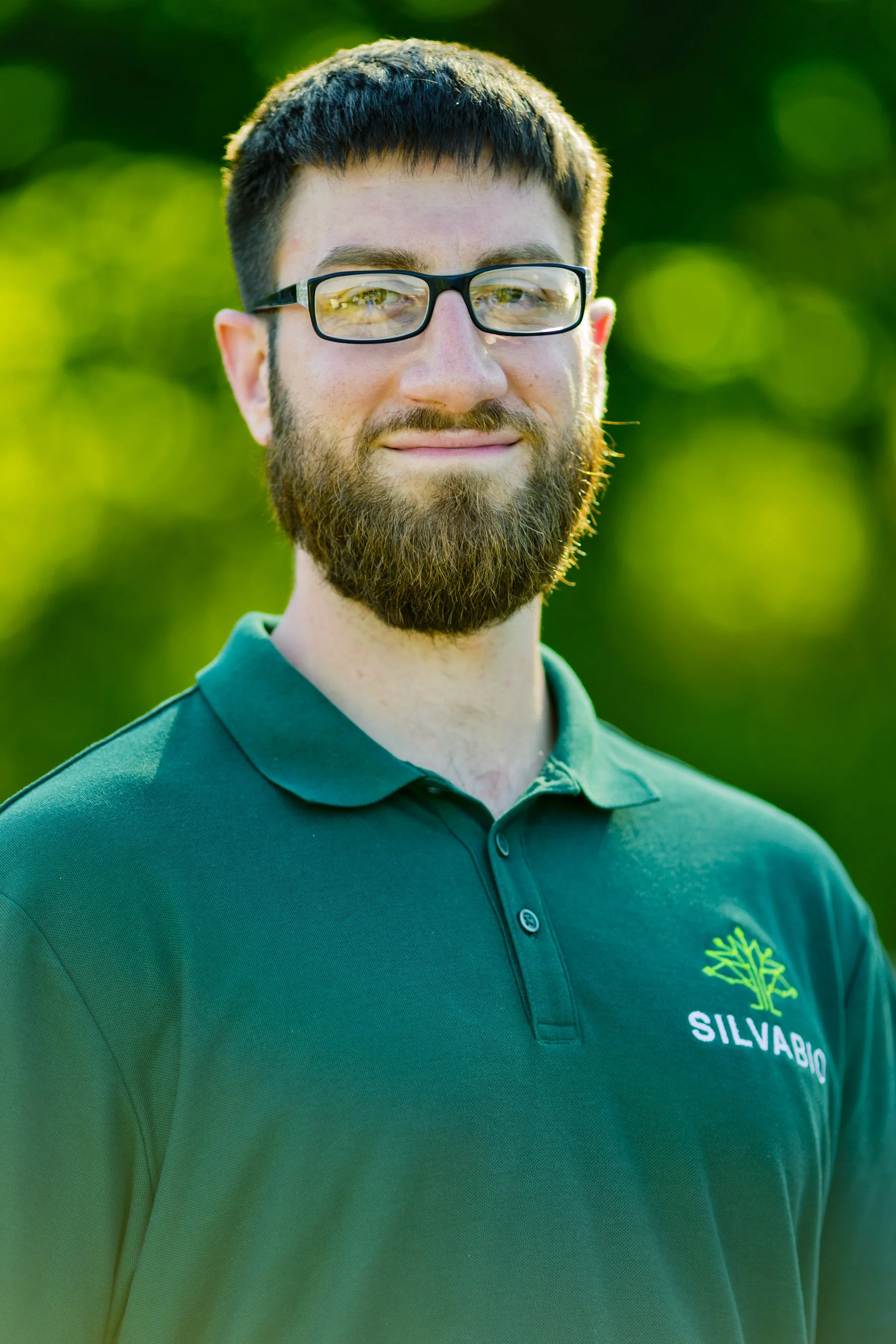 Man in a light purple polo shirt and black pants standing outdoors with greenery in the background.