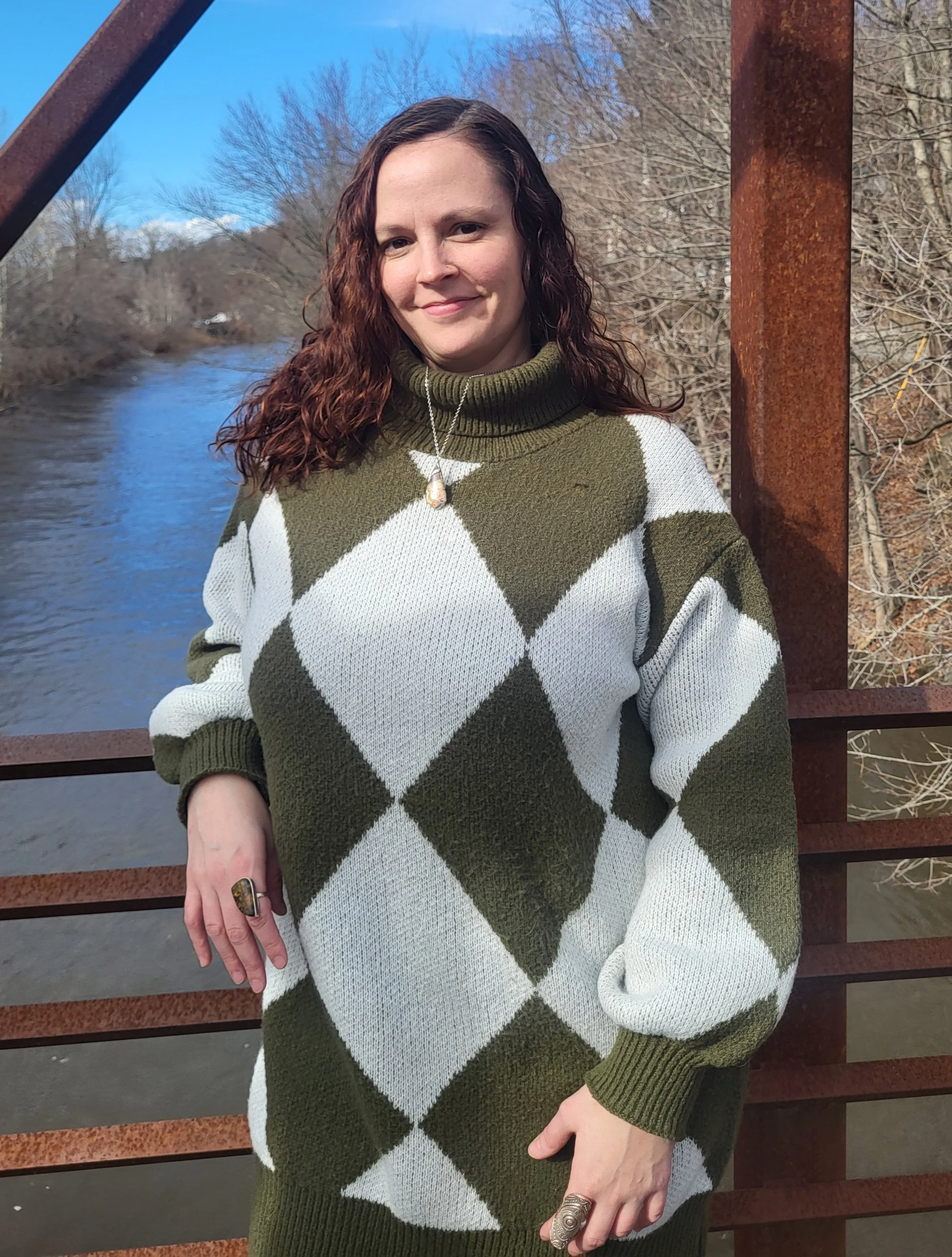 A woman with brown, curly hair standing on a bridge by a river, wearing a green and white argyle sweater, with trees and a blue sky in the background.