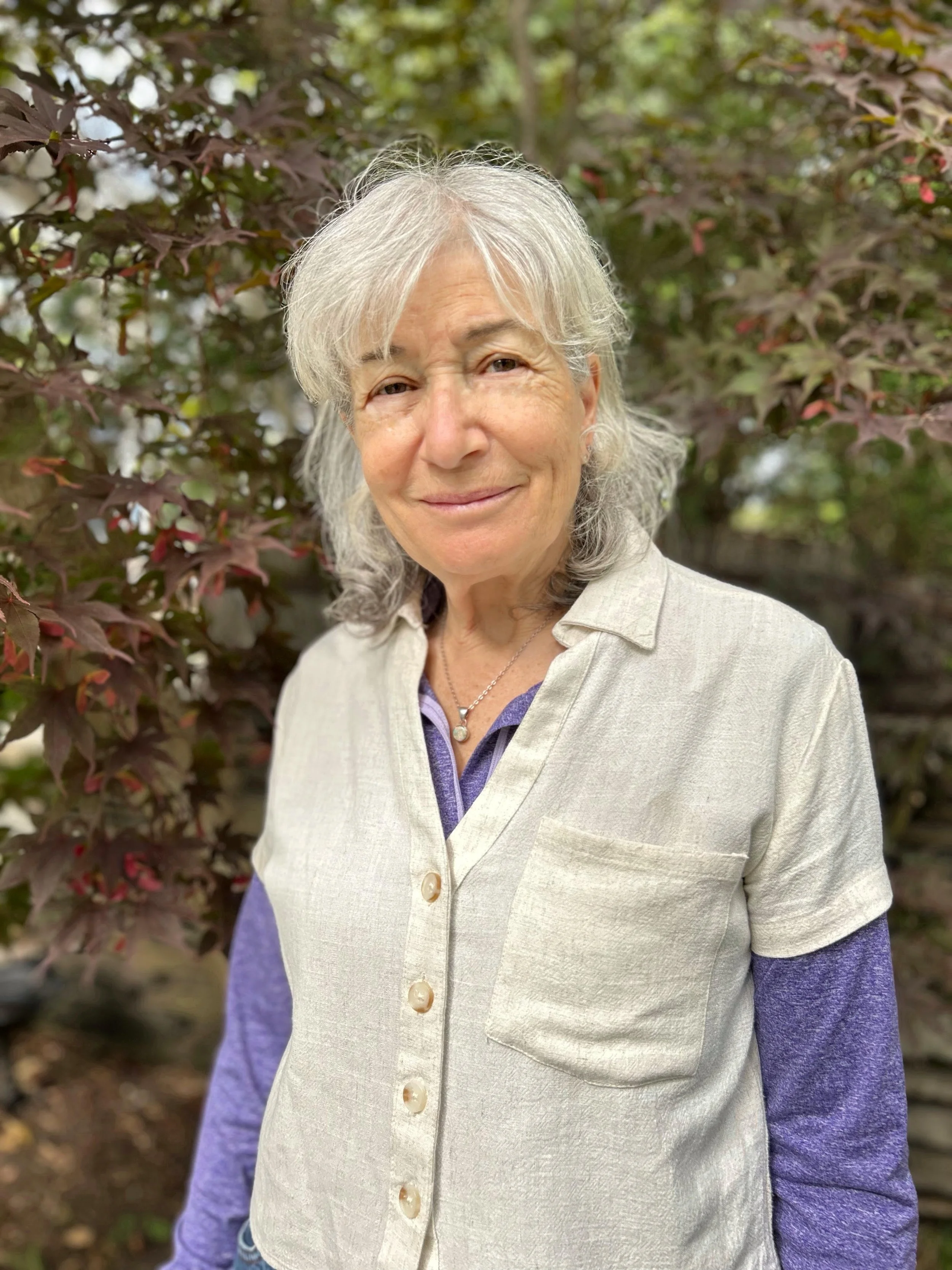 A smiling elderly woman with gray hair standing outdoors in front of leafy plants with reddish and green leaves. She wears a beige vest over a purple shirt and a necklace with a pearl pendant.