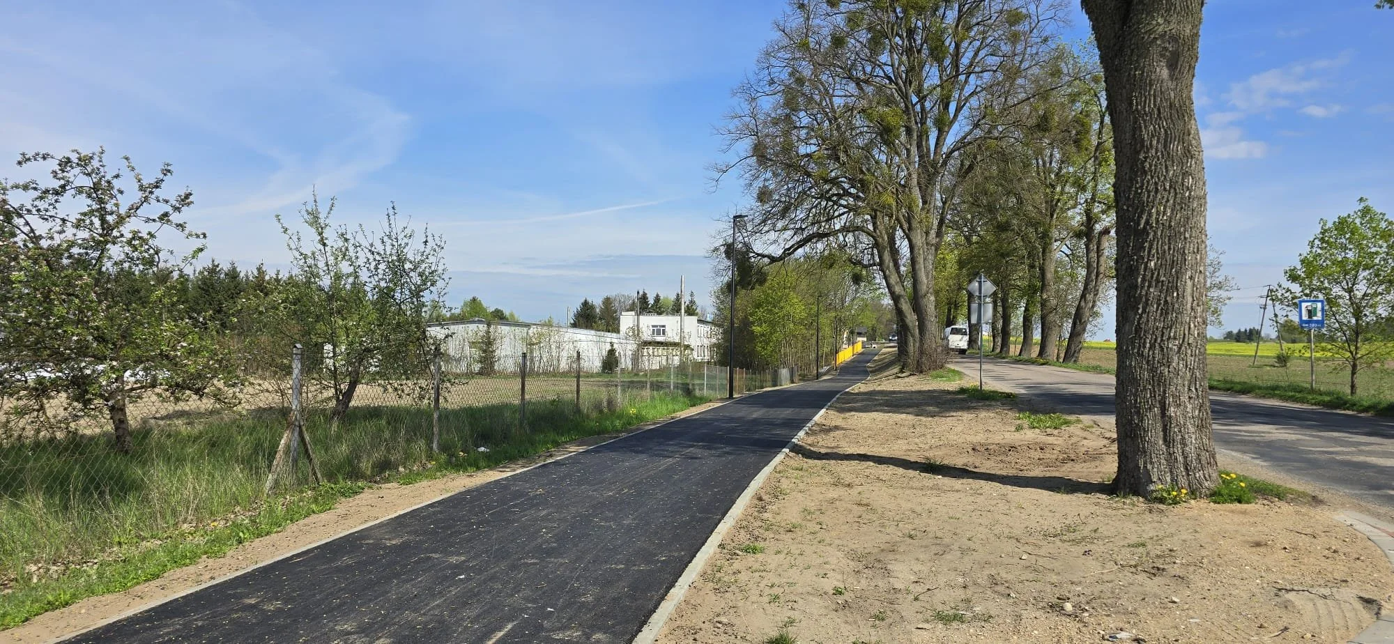 A newly paved sidewalk runs alongside a rural road with tall trees on one side and a fence with shrubs on the other. The sky is blue with some clouds, and there are white buildings and a van in the background.