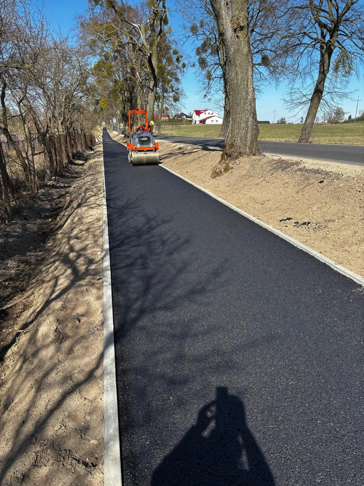 A person operating a vibratory roller compactor on a newly paved asphalt sidewalk in a rural area, with trees lining the sidewalk and a few houses in the background under a clear blue sky.