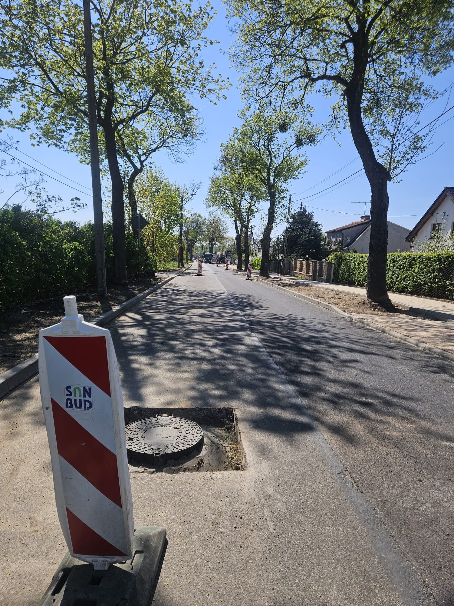 A street with trees on each side, some maintaining shadow over the road. There are construction barricades along the street, and a manhole cover in the foreground near a construction barrier. Residential houses are visible on the right side, and the 