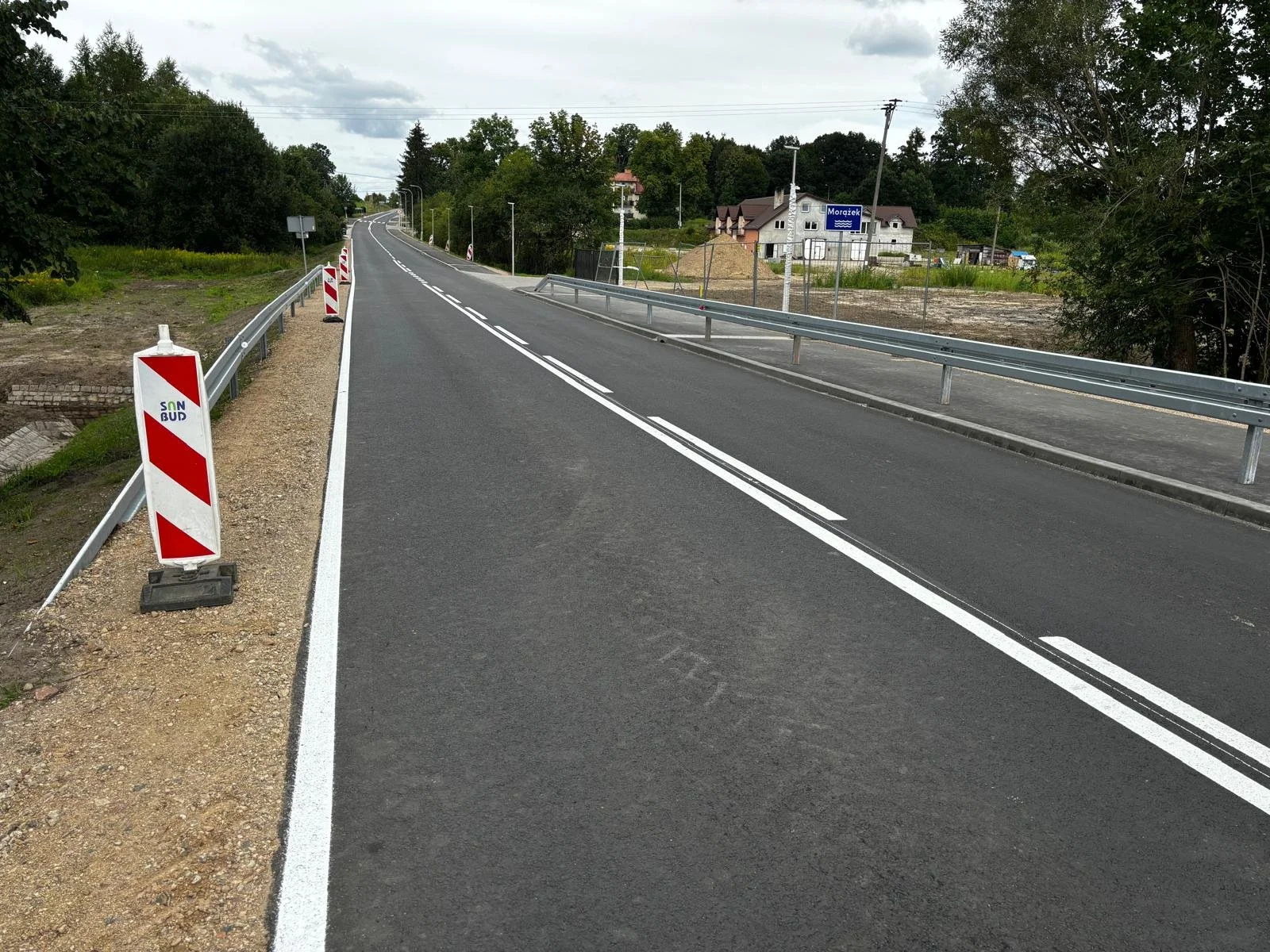 A newly paved road with white double dashed lines in the center, bordered by metal guardrails and construction barriers on the sides, in a rural area with trees, houses, and cloudy sky in the background.