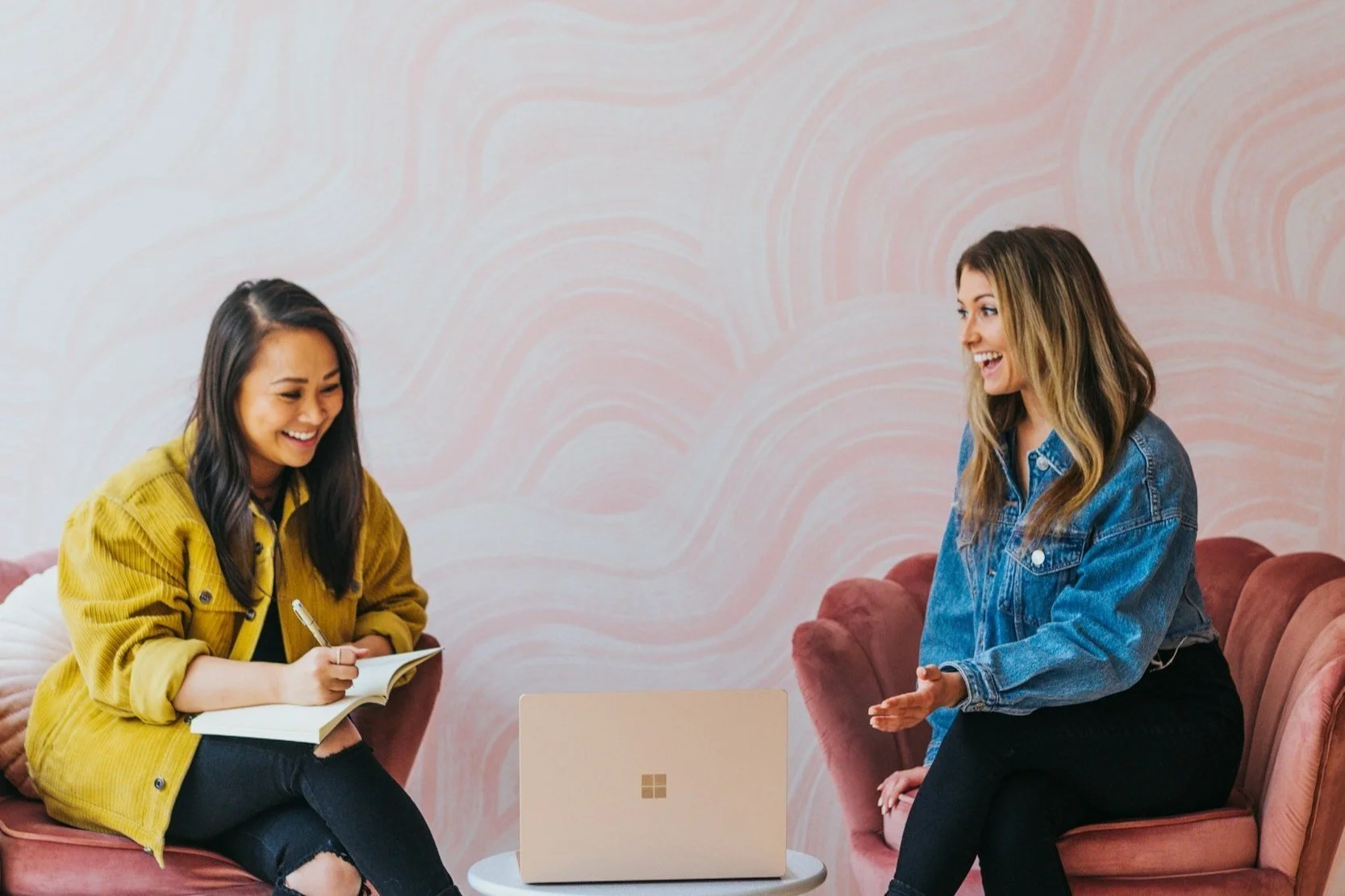 Two med spa owners sitting on pink couches, smiling and talking, with a beige laptop on a small white table between them and pink wavy-patterned wallpaper in the background.