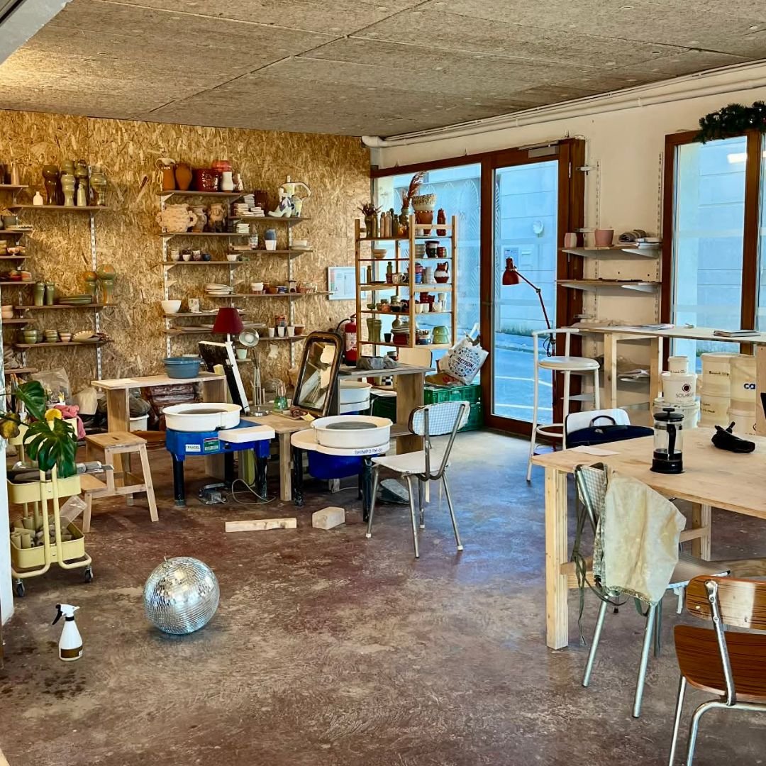 Un atelier avec des étagères remplies de céramiques et d'objets en argile, une table en bois avec des outils, une boule à facettes, et plusieurs chaises, fenêtres laissant entrer de la lumière naturelle. Atelier de céramique, lieu de cours 