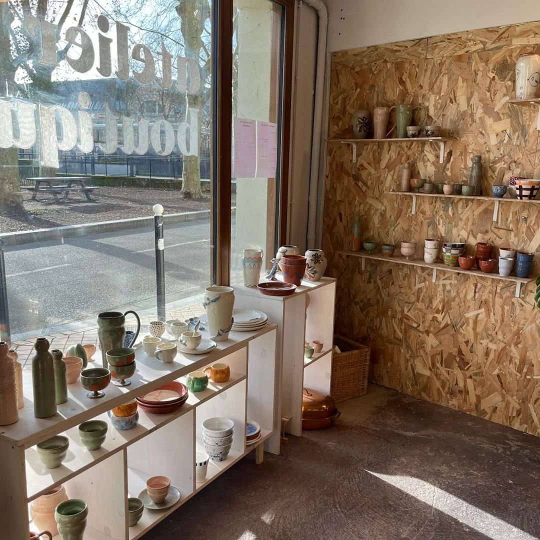 Une boutique de poterie avec des étagères en bois et des céramiques colorées exposées, à côté d'une fenêtre laissant passer la lumière naturelle.