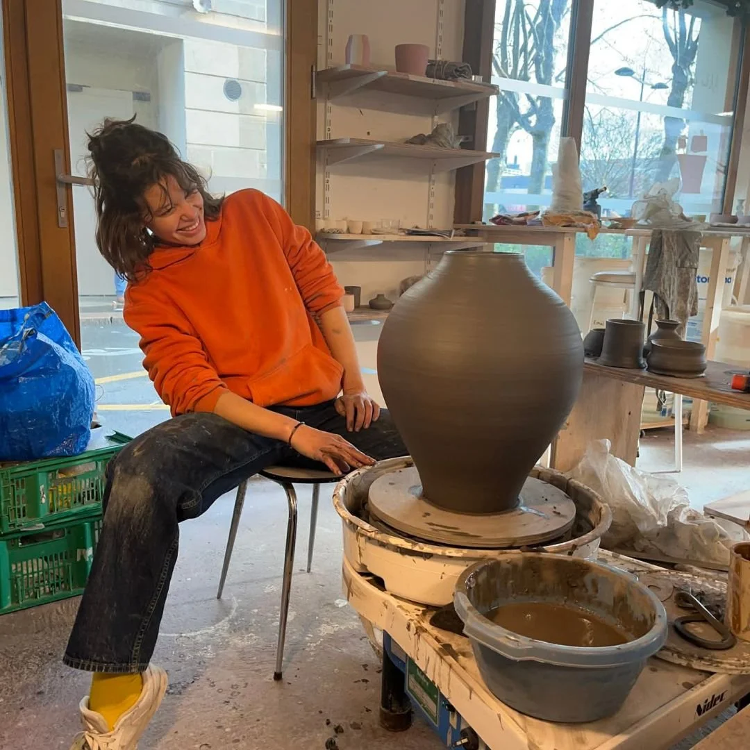 Une femme souriante en train de travailler sur un pot en argile sur un tour de potier dans un atelier de poterie, entourée de matériaux et d'outils de sculpture.