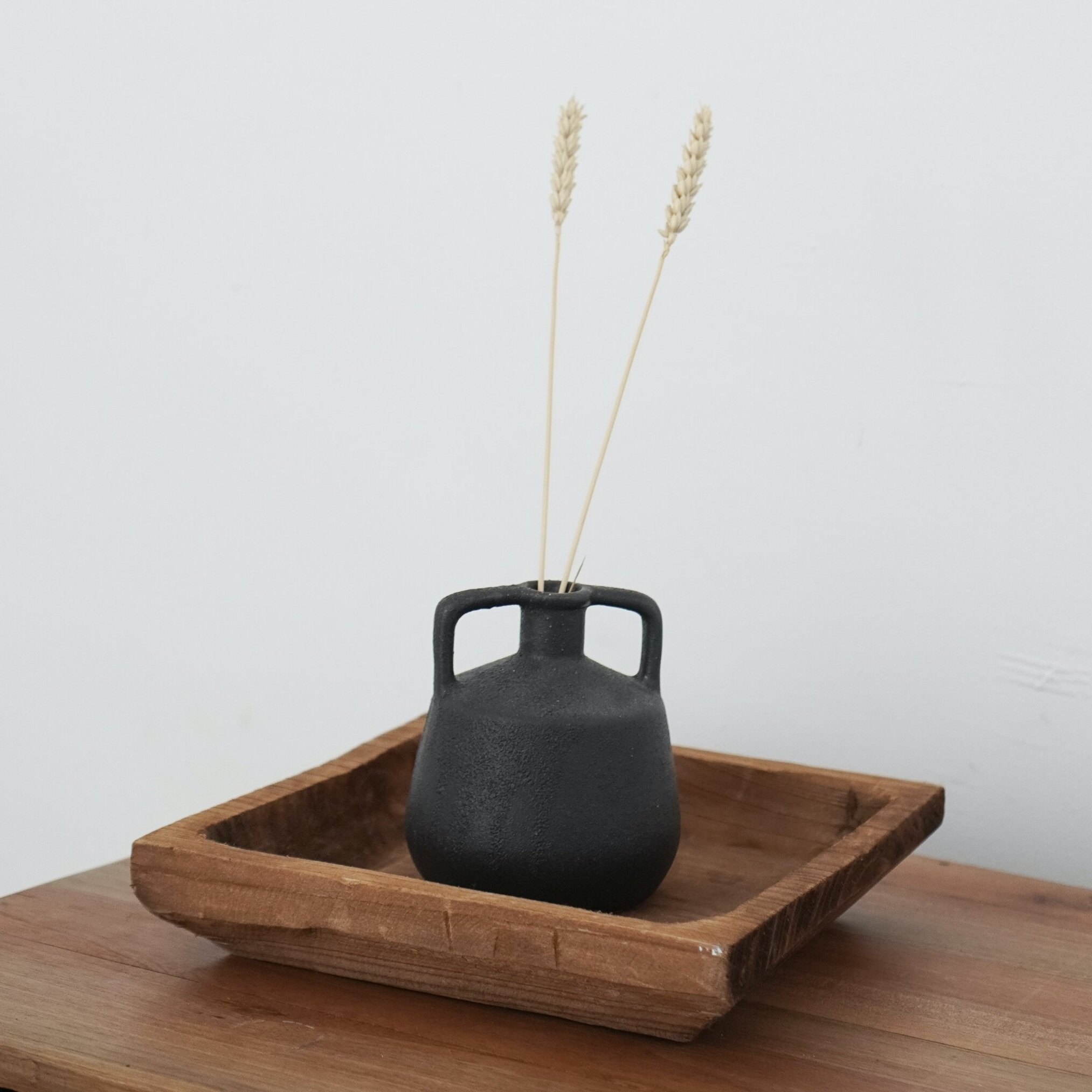 Black ceramic vase with two wheat stalks, placed inside a wooden tray on a wooden surface.