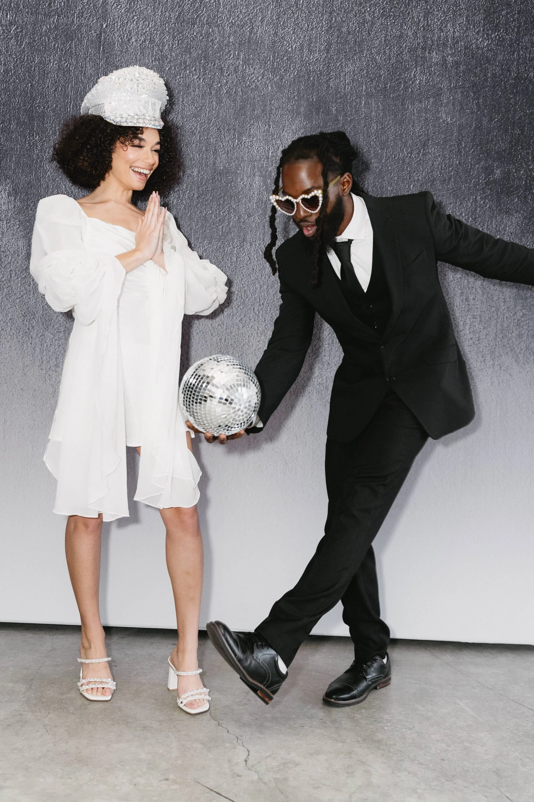 Two stylishly dressed individuals, a woman in a white dress and a man in a black suit with pearl-like glasses, celebrate with a disco ball against a dark wall.