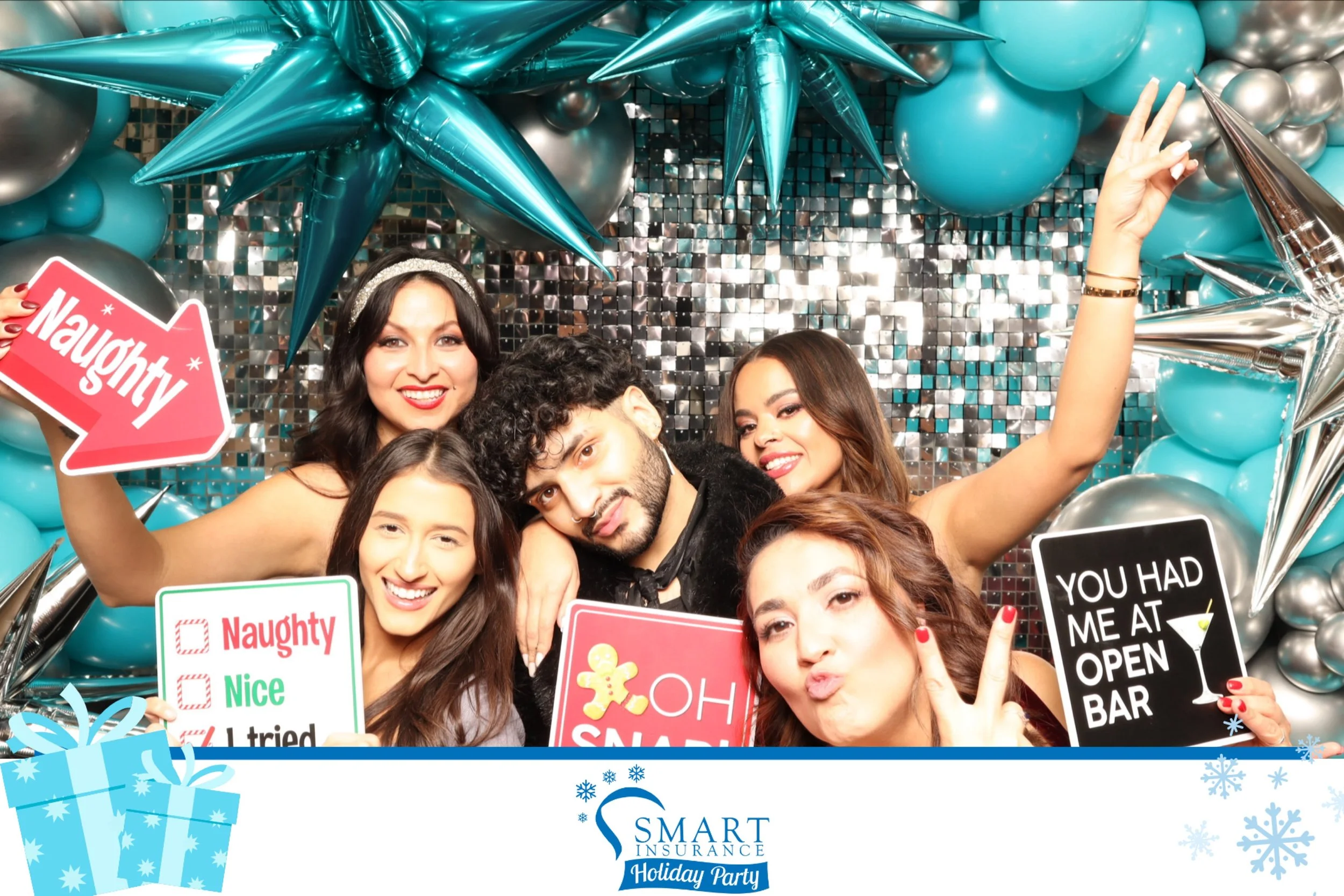 Group of five friends at a holiday party posing with party props against a shiny silver and blue balloon backdrop, with signs reading 'Naughty', 'You had me at open bar', and other playful messages.