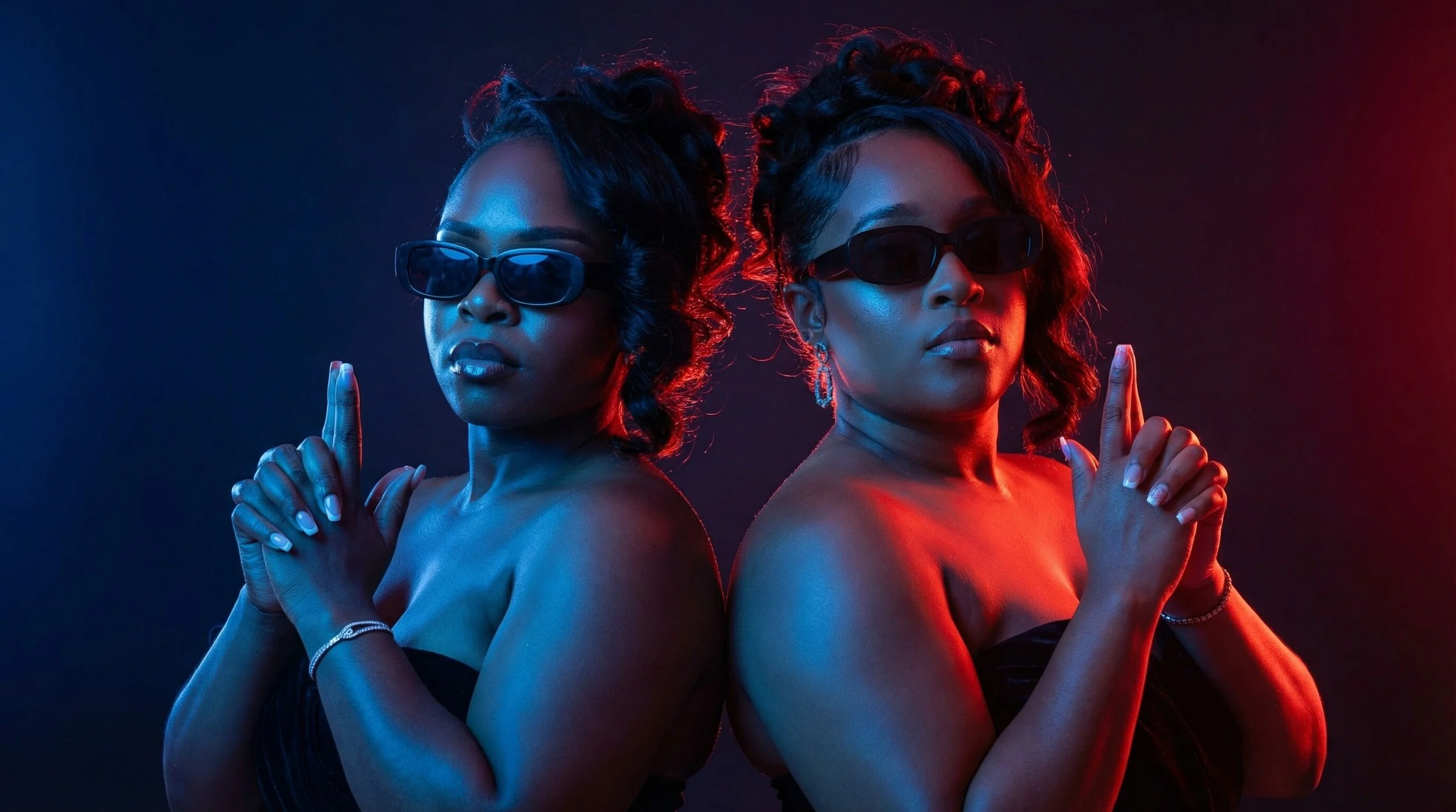 Two women with dark skin wearing black strapless dresses, black sunglasses, and jewelry, standing back-to-back with their hands together in a prayer pose, illuminated by blue and red neon lights.