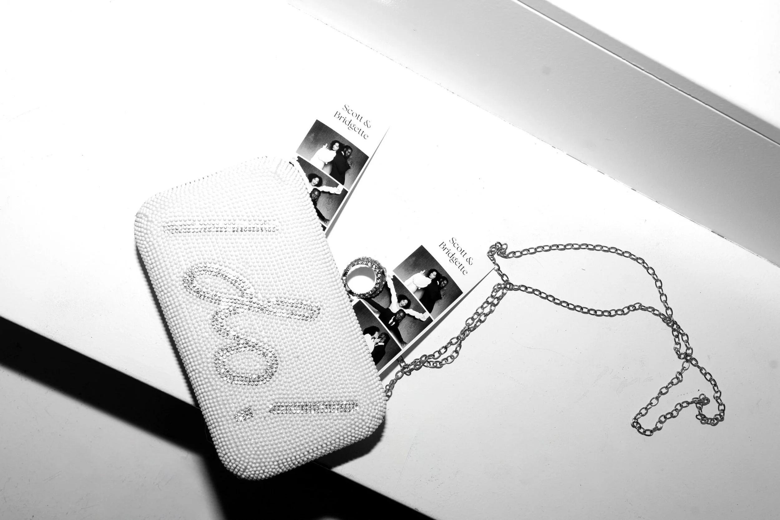 A white beaded clutch with the Chanel logo, a chain necklace, and two photo booth strips of a woman and man, placed on a white surface.