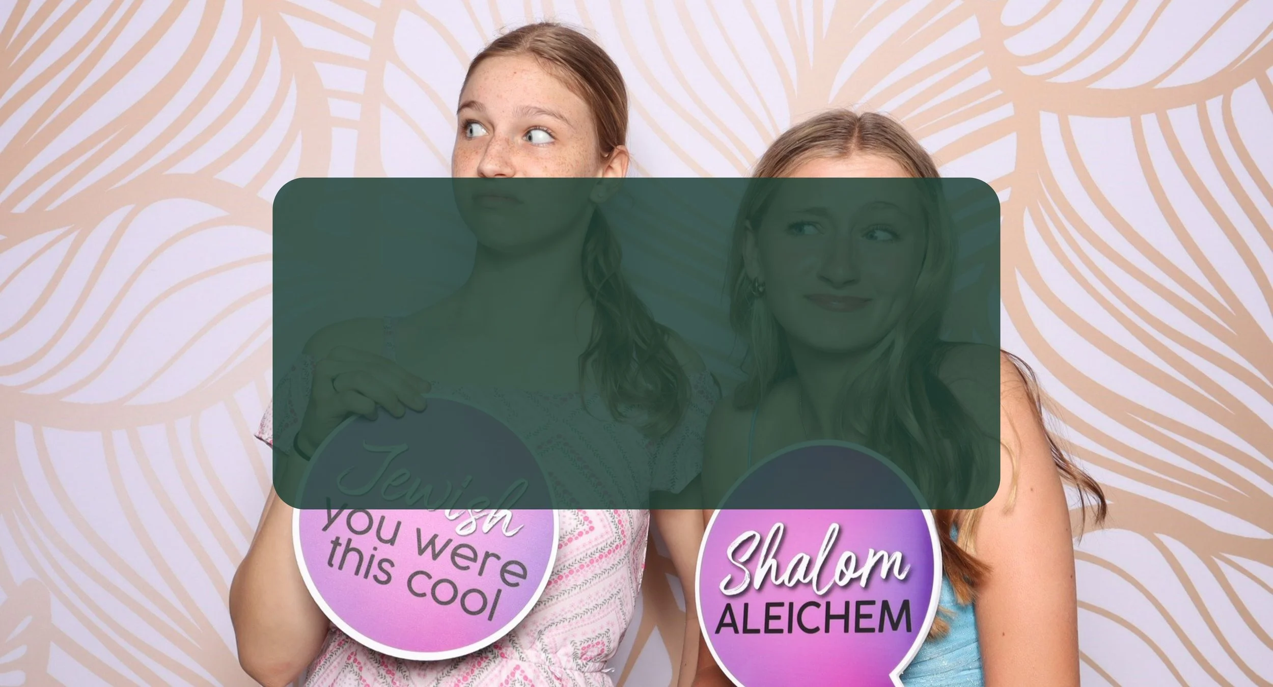 Two young girls holding circular signs with Hebrew and English text, standing against a patterned backdrop.