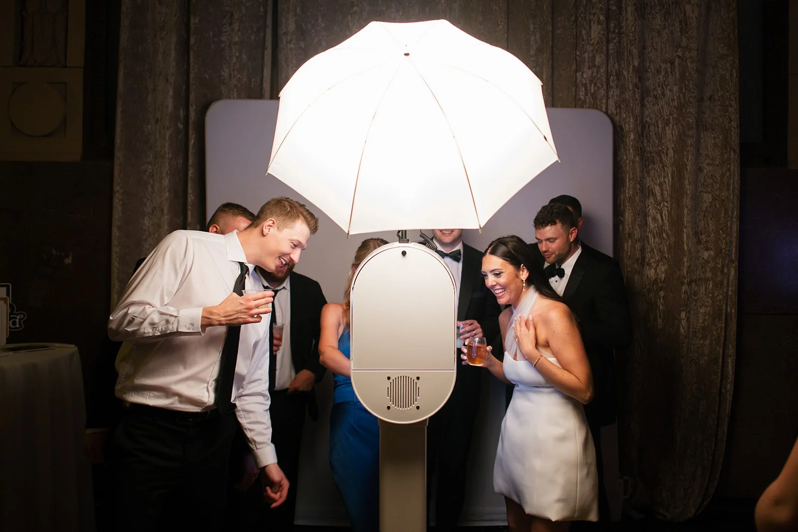 Group of people at a photo booth, laughing and holding drinks, under a large umbrella-shaped light.