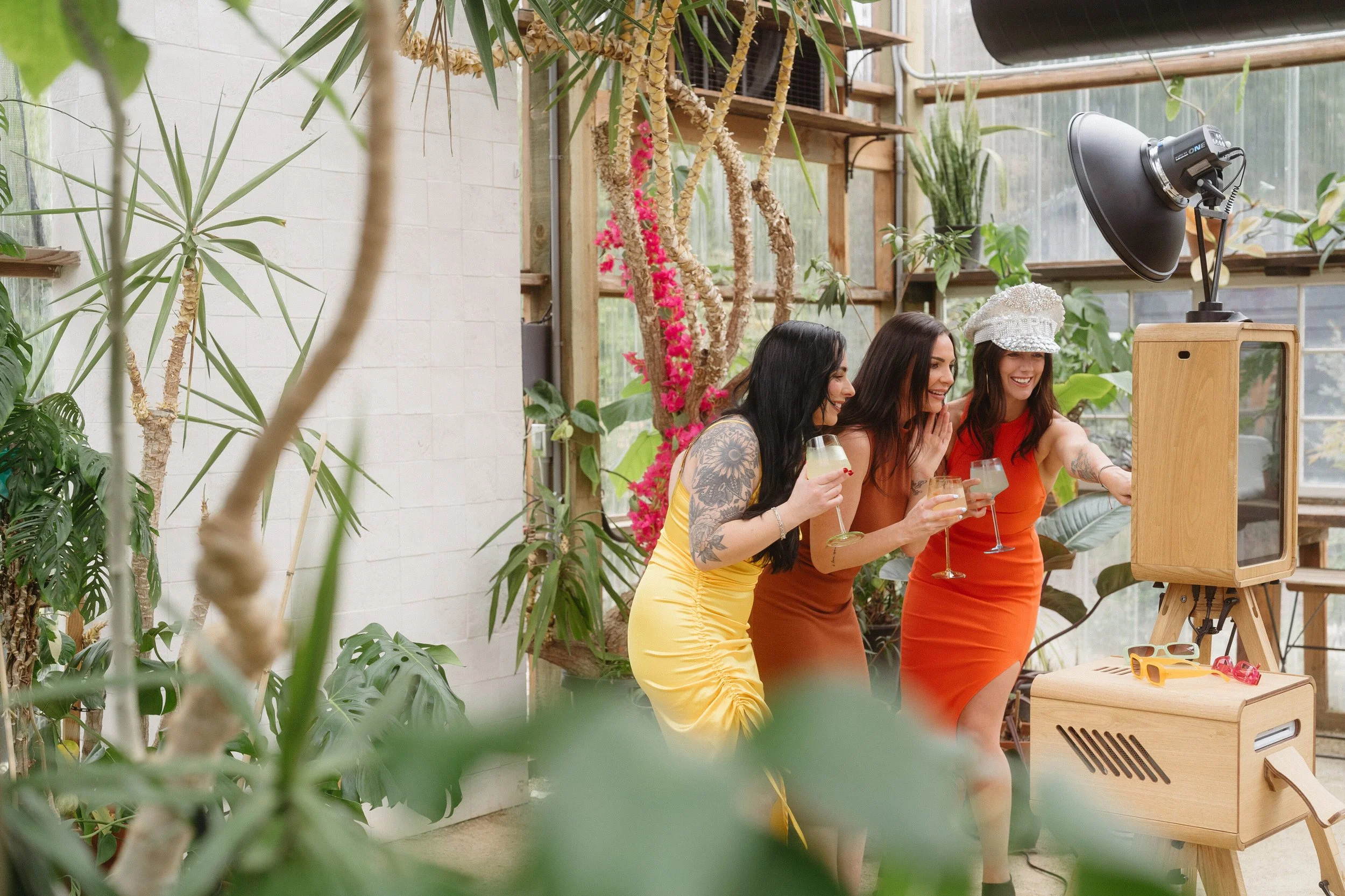 Three women at a party are looking at a vintage photo booth inside a greenhouse filled with plants. They are smiling and holding drinks, with one woman wearing a white hat and an orange dress, another in a yellow dress, and the third in a rust-colored dress.
