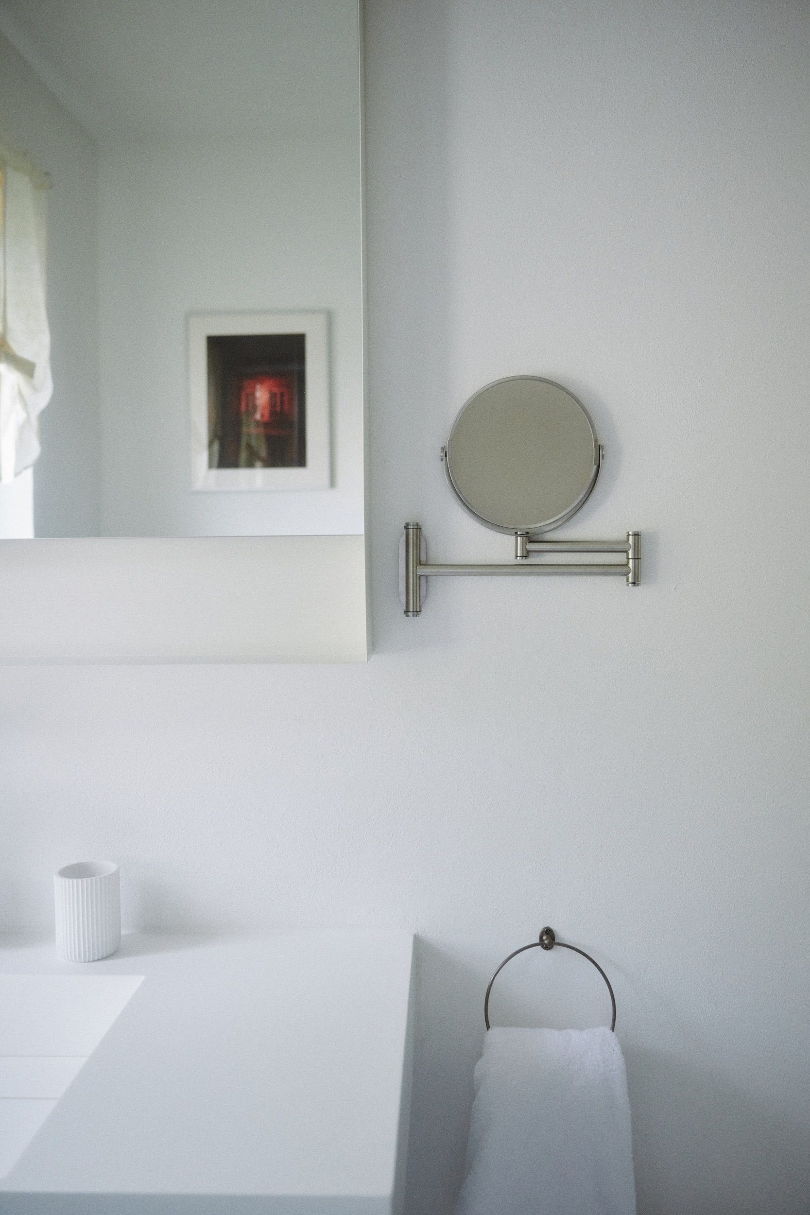 Ein Badezimmer mit einem runden, schwenkbaren Spiegel an der Wand, einem weißen Waschbecken, einem Handtuchhalter mit einem weißen Handtuch und einem kleinen, geriffelten Behälter. Im Hintergrund ist ein Fenster mit Vorhang sichtbar.