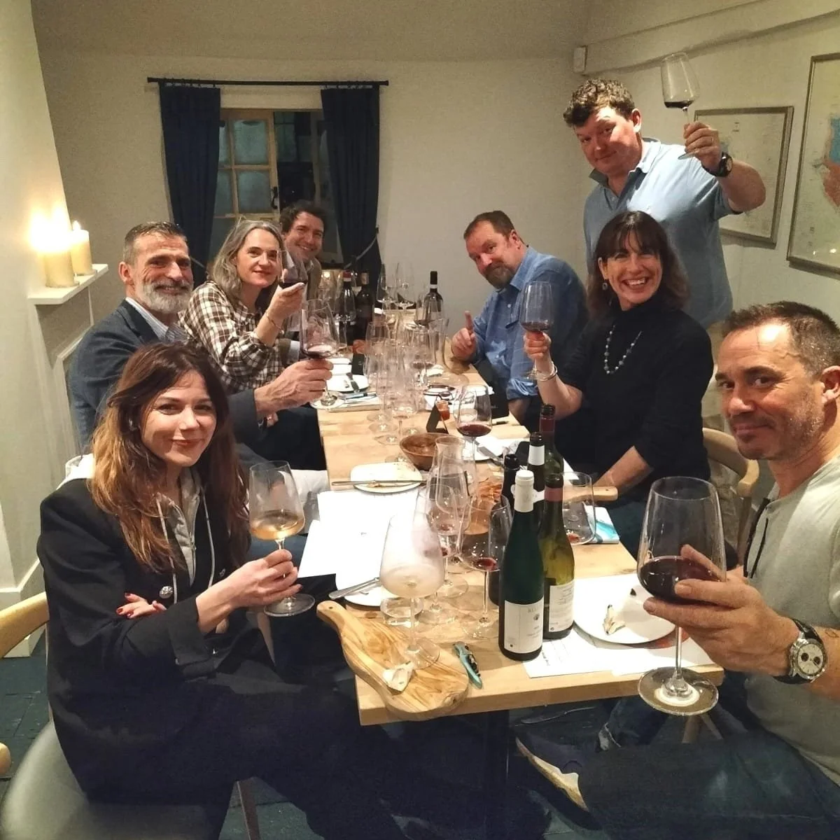 A group of nine people sitting and standing around a dining table, raising glasses of red wine in a cozy, well-lit room.