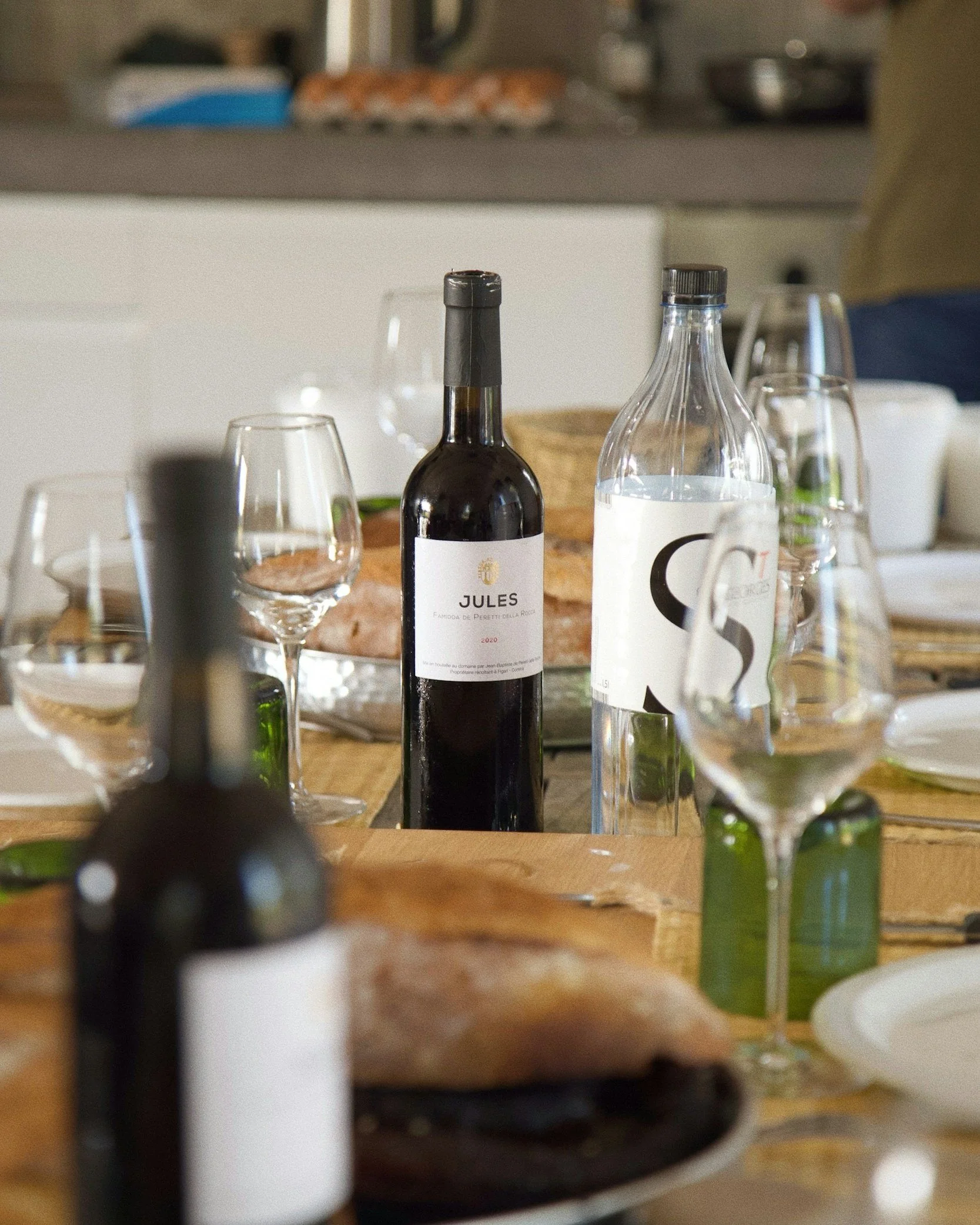 Table set for a meal with a bottle of red wine, glasses, a bottle of water, and a loaf of bread with a baked dish in the background.