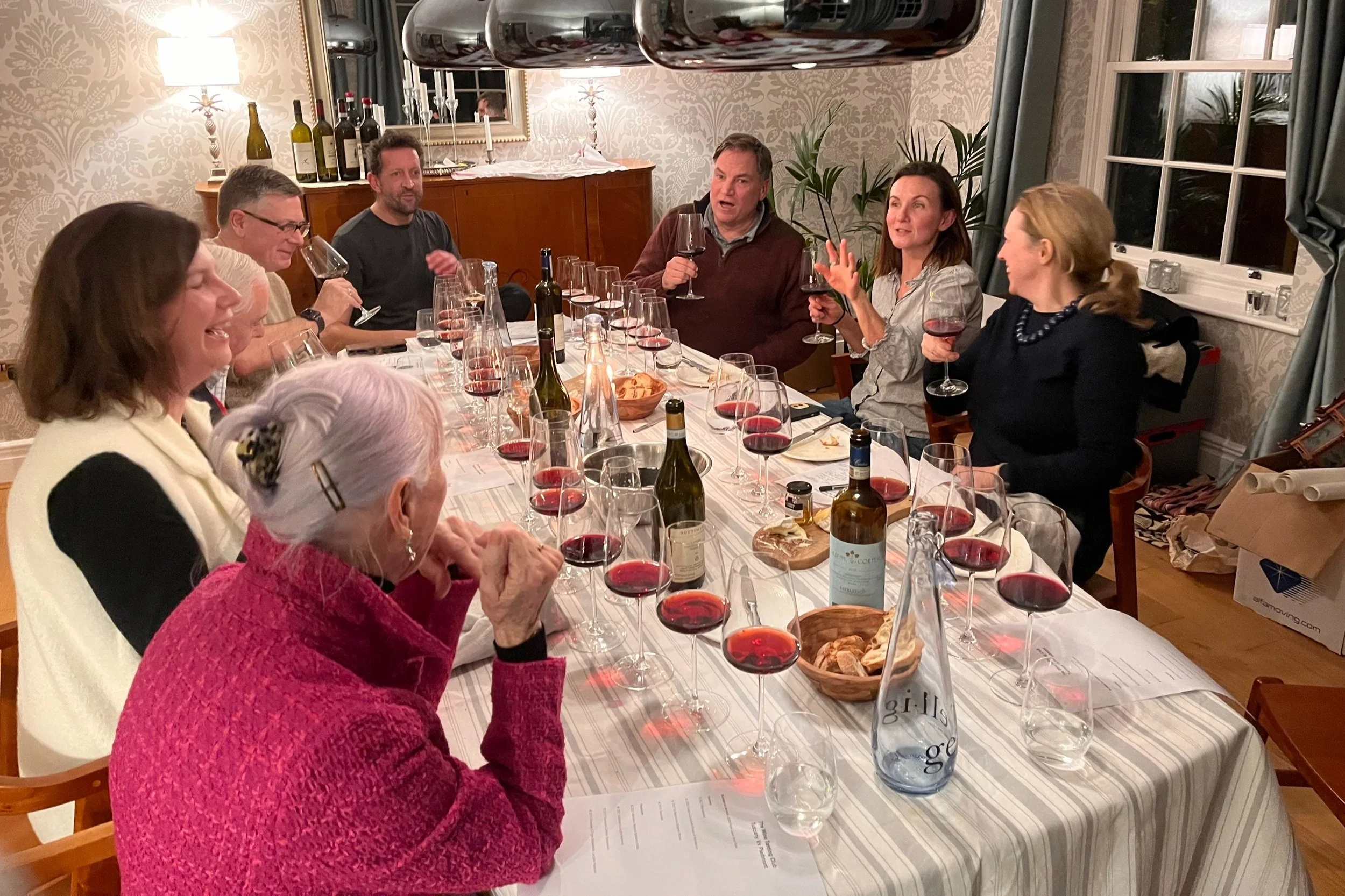 Group of people sitting around a dining table enjoying a wine tasting, with several glasses of red wine, bottles of wine, and snacks on the table, in a warmly decorated room.