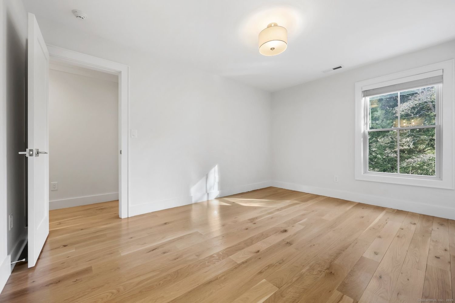 Empty room with hardwood floor, white walls, a window with a view of greenery, and a ceiling light fixture.