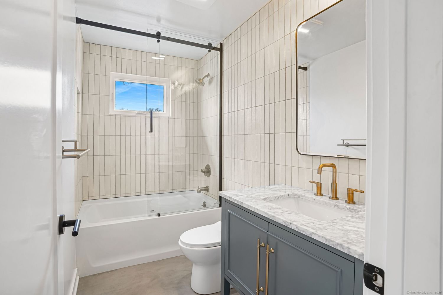 Modern bathroom with white tiled walls, bathtub with glass door, toilet, gray vanity with marble countertop, gold faucets, and a rectangular mirror.
