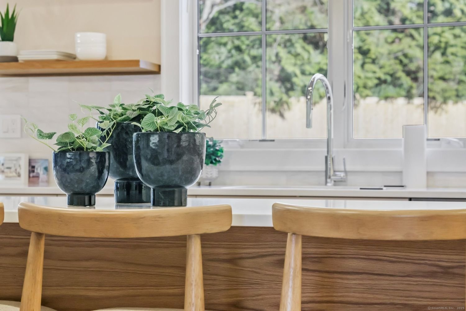 Modern kitchen with potted plants on counter, wooden chairs, floating shelf with dishes, and sink with window view.