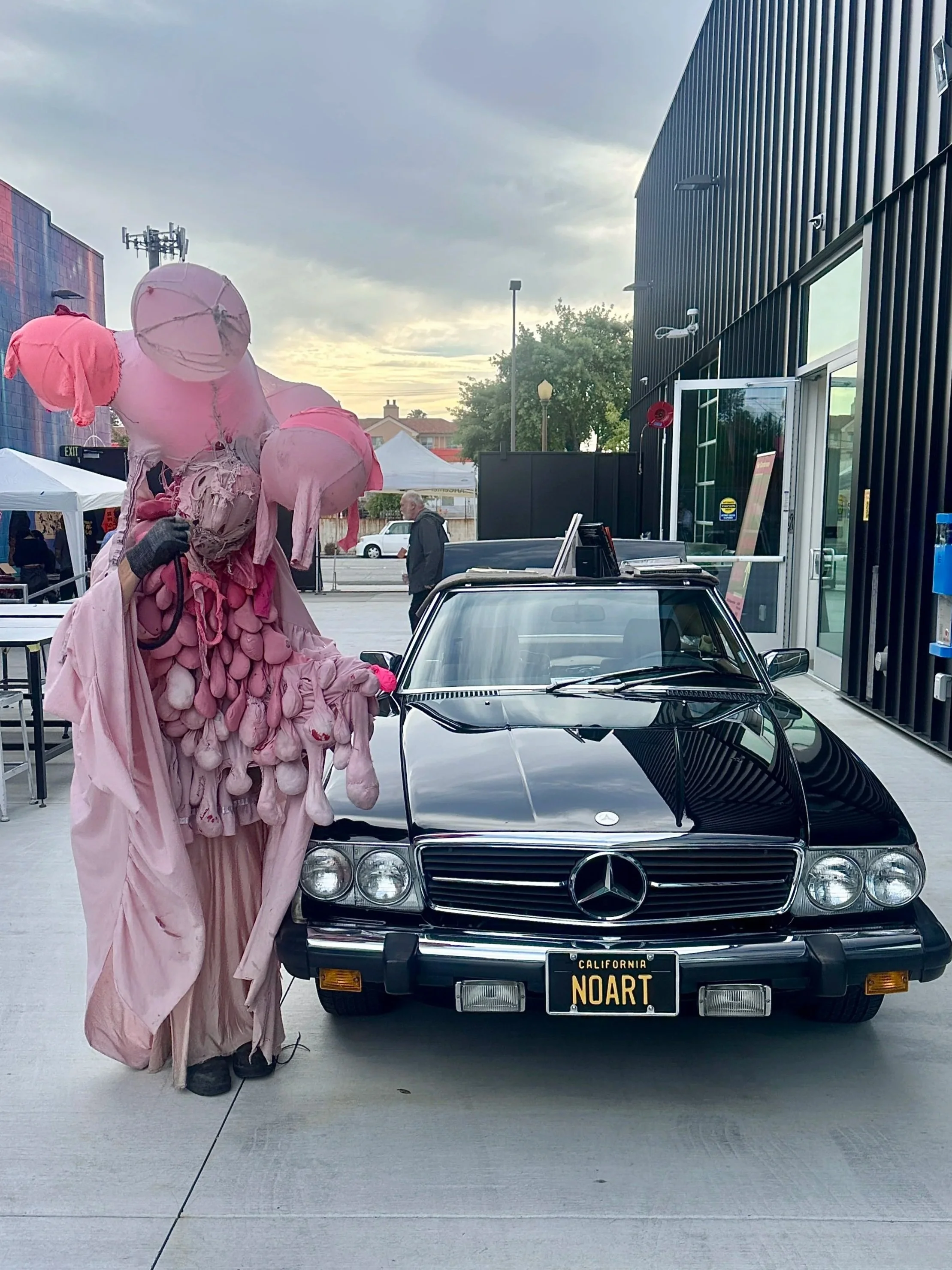 A person in an elaborate pink costume with a large pink headpiece, standing next to a black vintage Mercedes-Benz car with California license plates that reads 'NOART.' The scene appears to be outdoors near a modern building with a black exterior, in