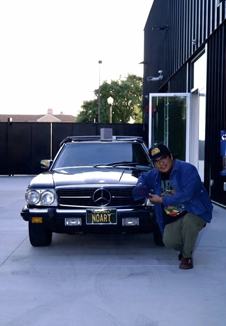 A person crouching next to a black Mercedes-Benz car with a customized California license plate that reads 'NOART'. The person is wearing glasses, a black cap, a blue jacket, and khakis, and is holding a cap. The setting appears to be outside a moder