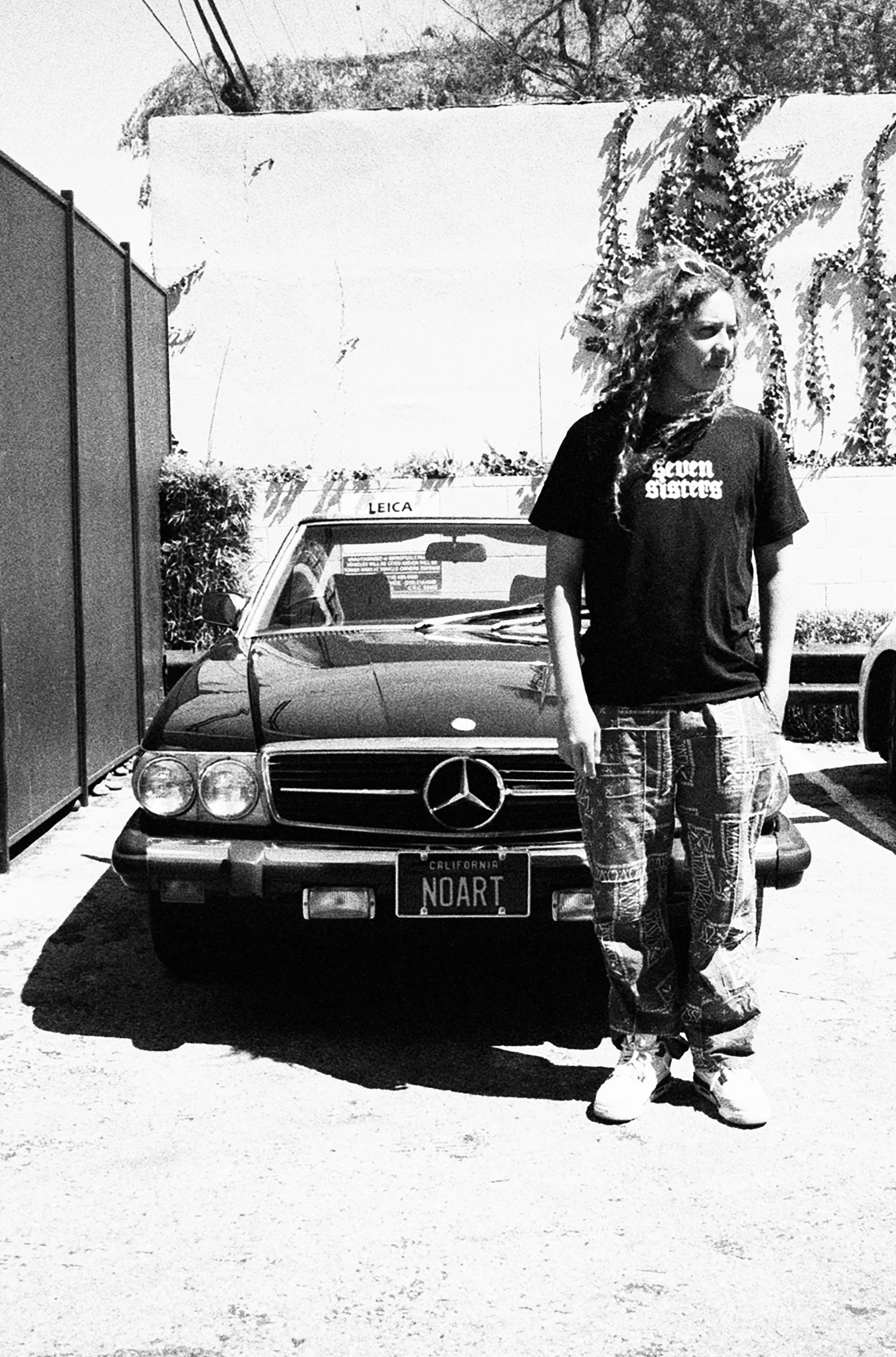 A person with long curly hair standing in front of a vintage Mercedes-Benz car with a California license plate reading "NOART". The person is wearing a black t-shirt, patterned pants, and sneakers. The person is also wearing a bracelet and appears to