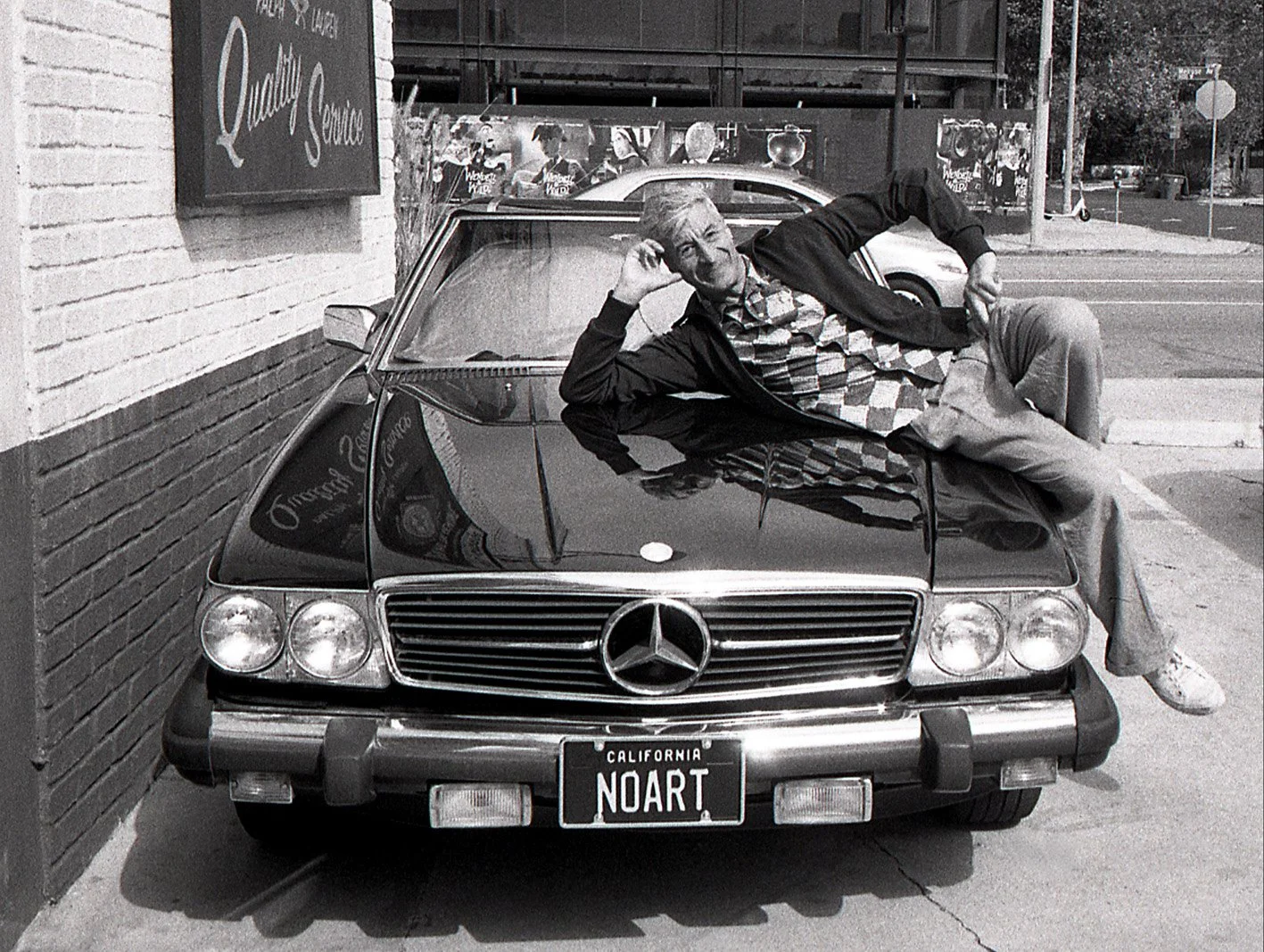 A man lying on the hood of a vintage black Mercedes-Benz car, with a California license plate reading 'NOART', parked on a city sidewalk next to a brick building and a street with traffic signs in the background.