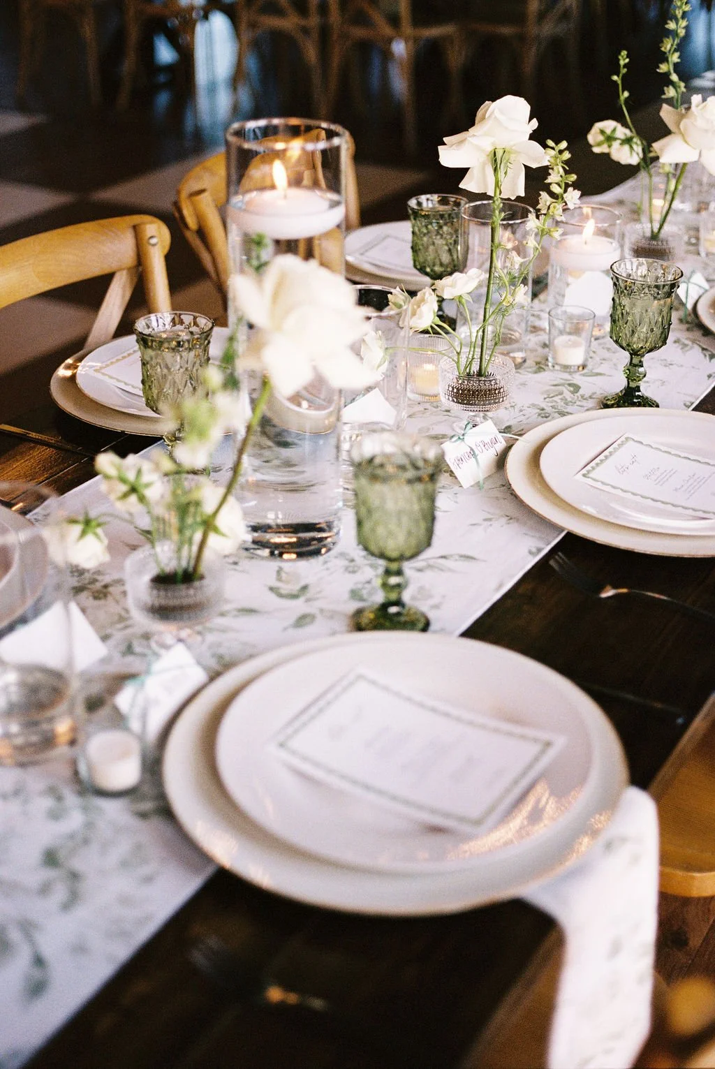 Wedding reception inside of River Road Venue, with dark wooden farm tables draped with floral patterned runners, an all white flowers in glass bud vases