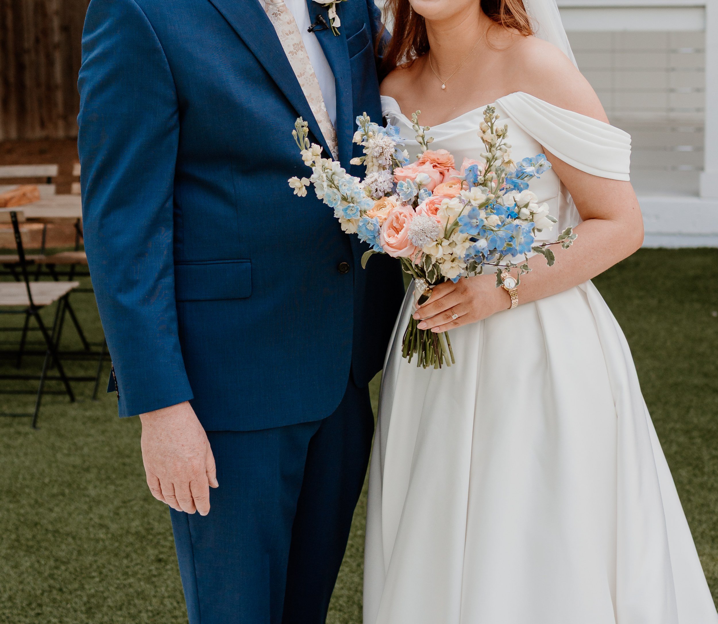 Bride holding a pastel bouquet next to a man in a blue suit with a boutonniere, white building behind (flower types unsure)