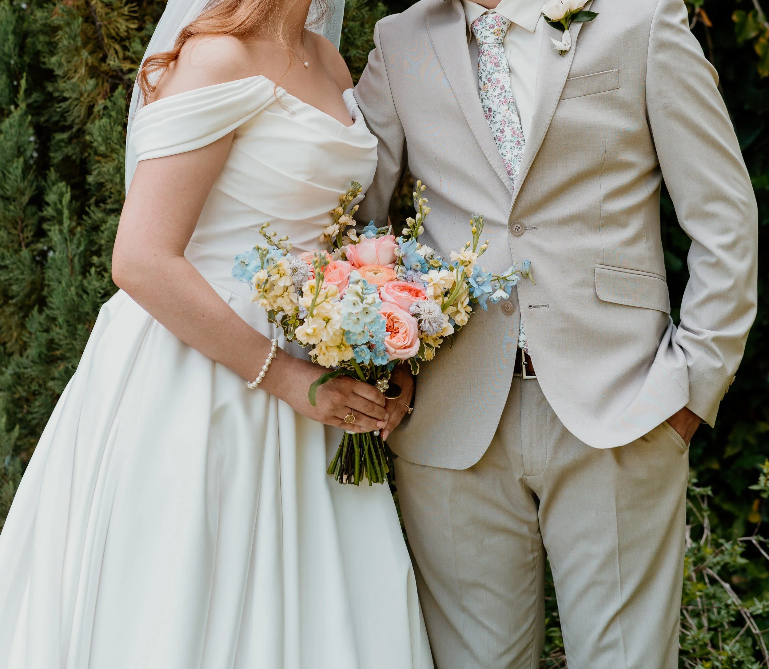 Couple outdoors by greenery, the bride holding a pastel bouquet of peach, pink and pale blue flowers and the groom wearing a small boutonniere (flower types unsure)