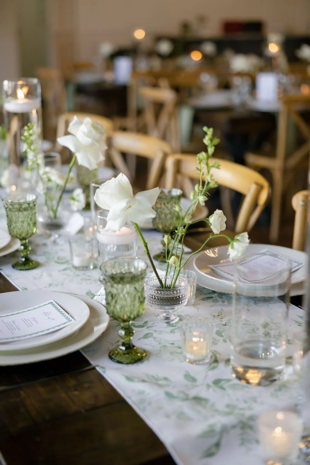 Wedding reception inside of River Road Venue, with dark wooden farm tables draped with floral patterned runners, an all white flowers in glass bud vases