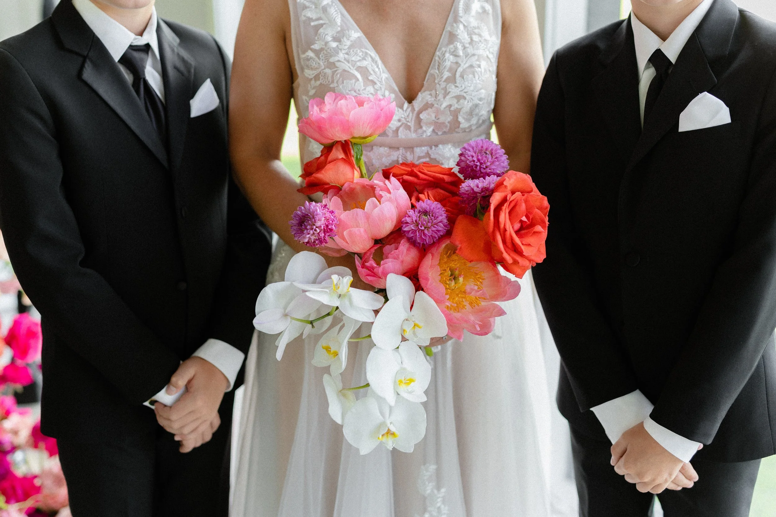 Bride holding a pink, coral and white orchid bouquet between two groomsmen