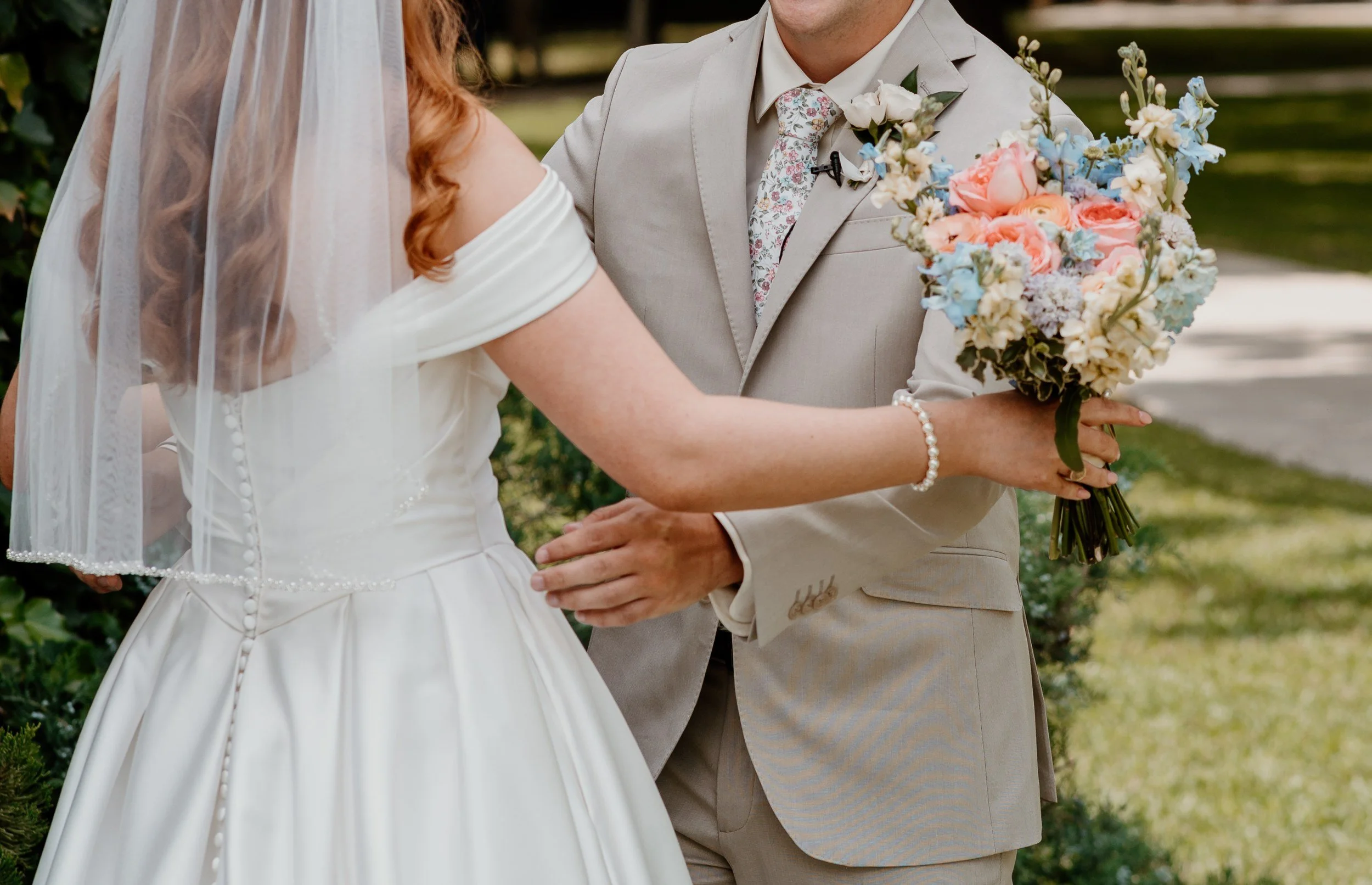 Groom laughing as he holds a pastel bouquet of peach, pink and blue flowers, the bride facing him in her veil (flower types unsure)