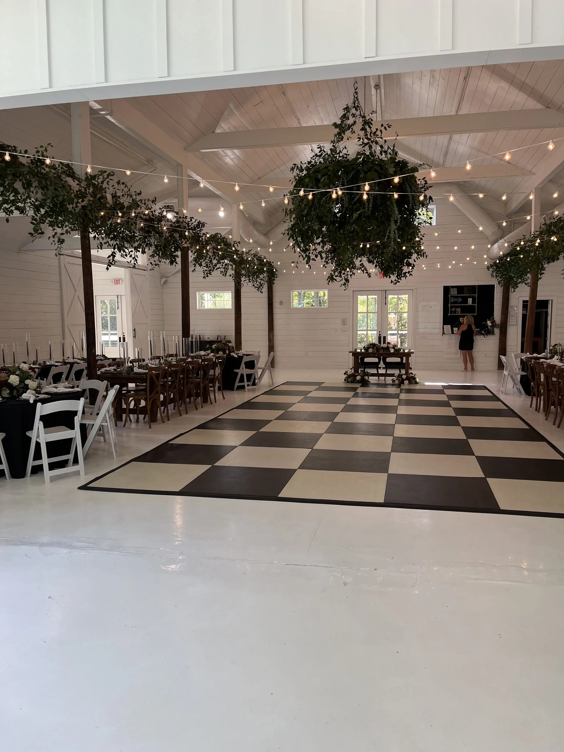 Open white barn reception hall with checkered dance floor, string lights and greenery by Haute Poppies