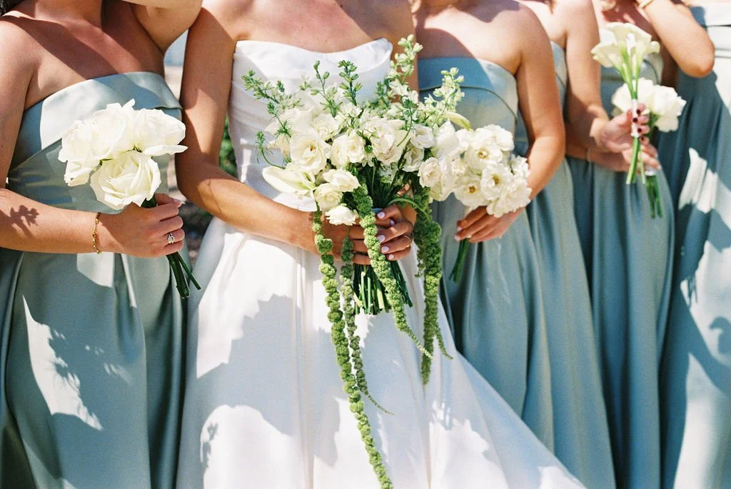 A detail of a bridal bouquet with white garden roses, lisianthus, and trailing green hanging amaranthus, and the bridesmaids holding single flower bouquets of roses, garden roses, calla lillies