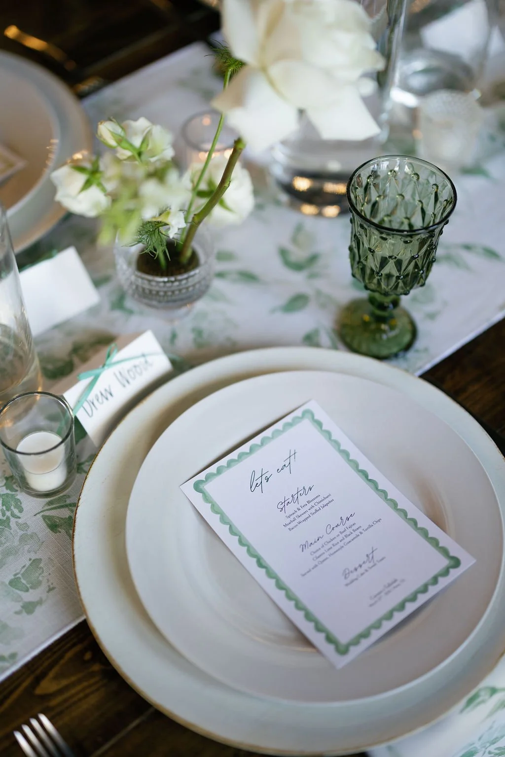 Details of white plates with a square menu on top, with scallop details, greenery patterned table runners, and bud vases by Haute Poppies Floral