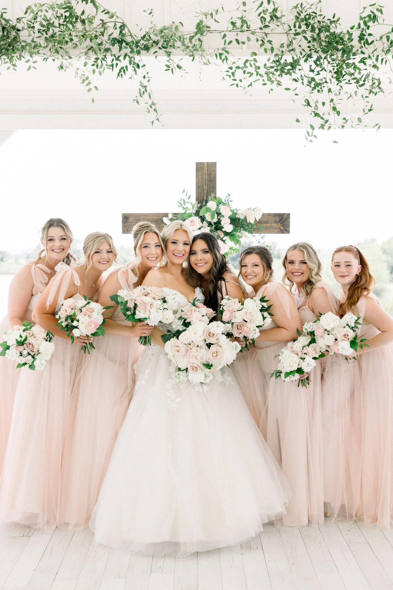 Bride and bridesmaids in blush gowns holding white and blush bouquets in front of a wooden ceremony cross with one white and blush floral pod