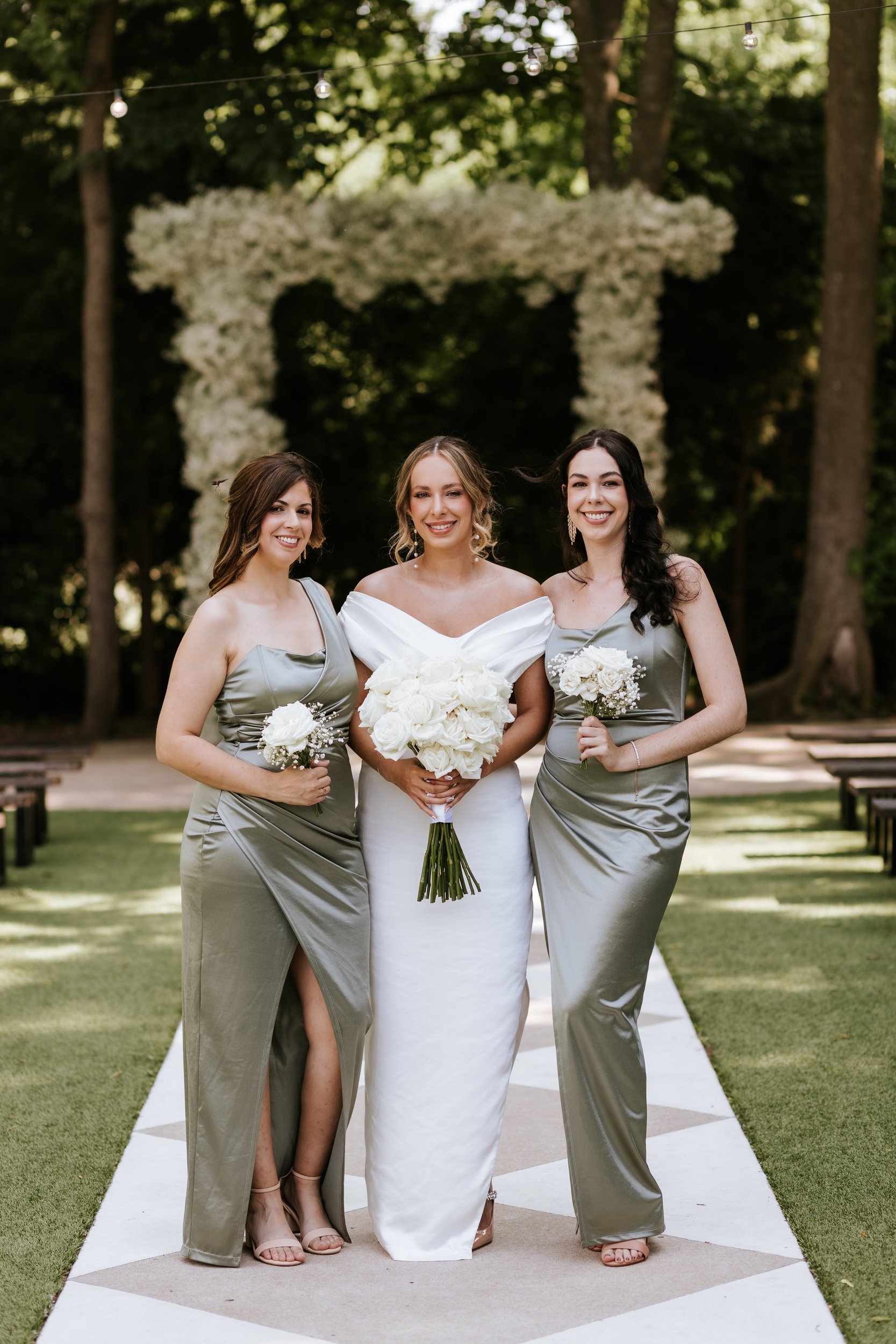 Bride with bridesmaids in sage holding white bouquets on a checkered aisle, white floral arbor behind them