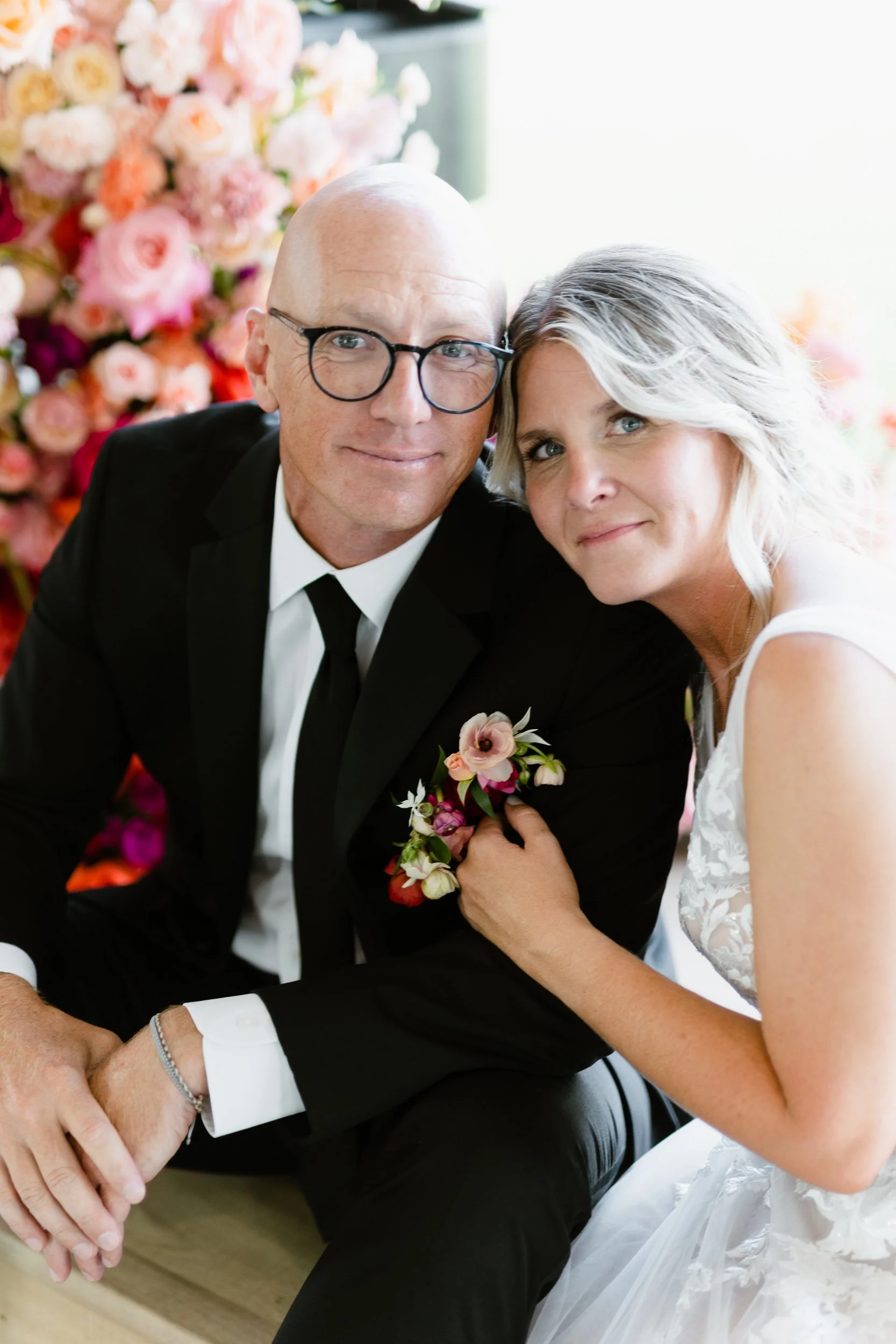 Couple smiling close together, groom wearing a pink and red boutonniere, floral install behind