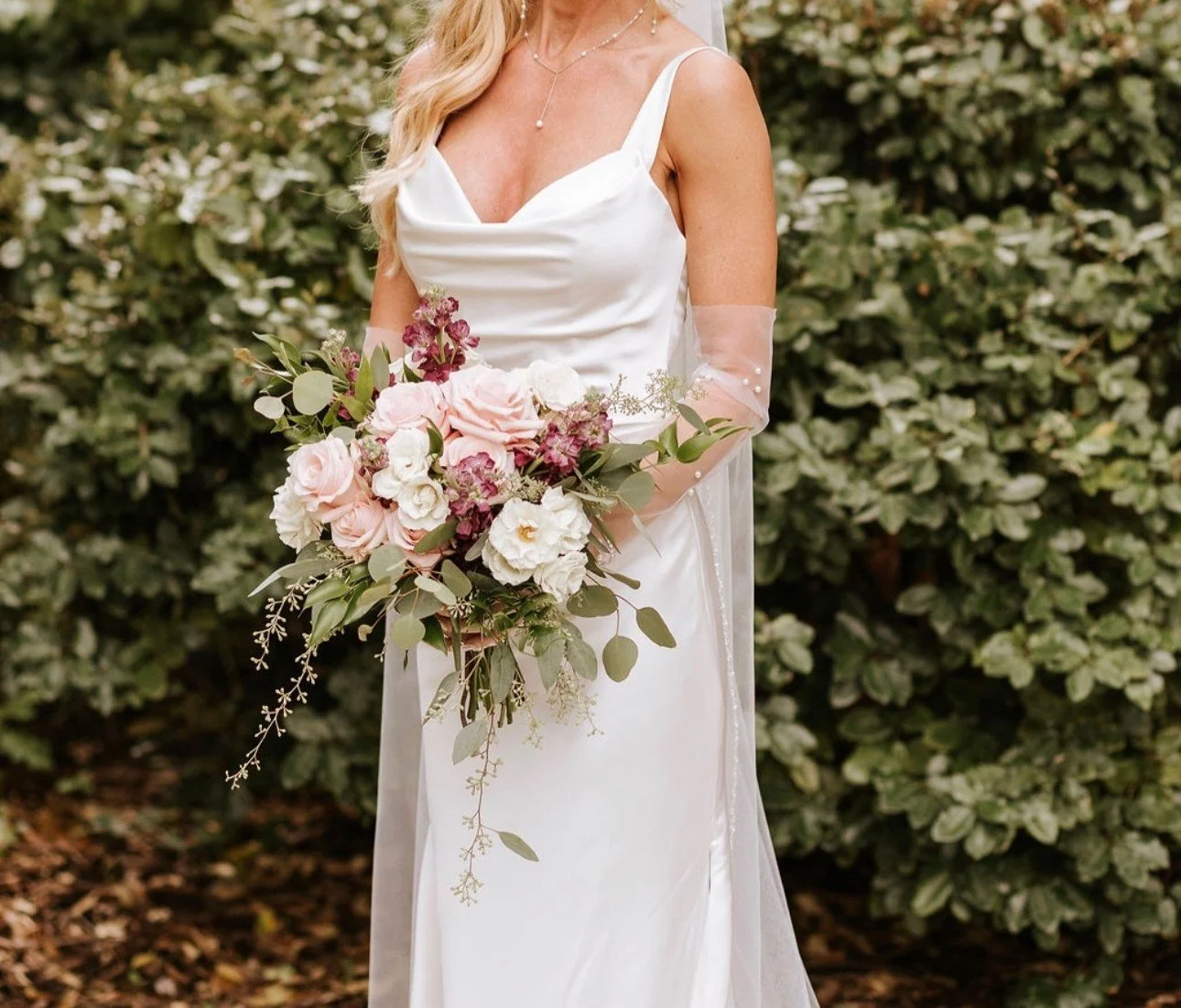 Bride in a garden holding a blush, white and burgundy bouquet with trailing greenery (flower types unsure)