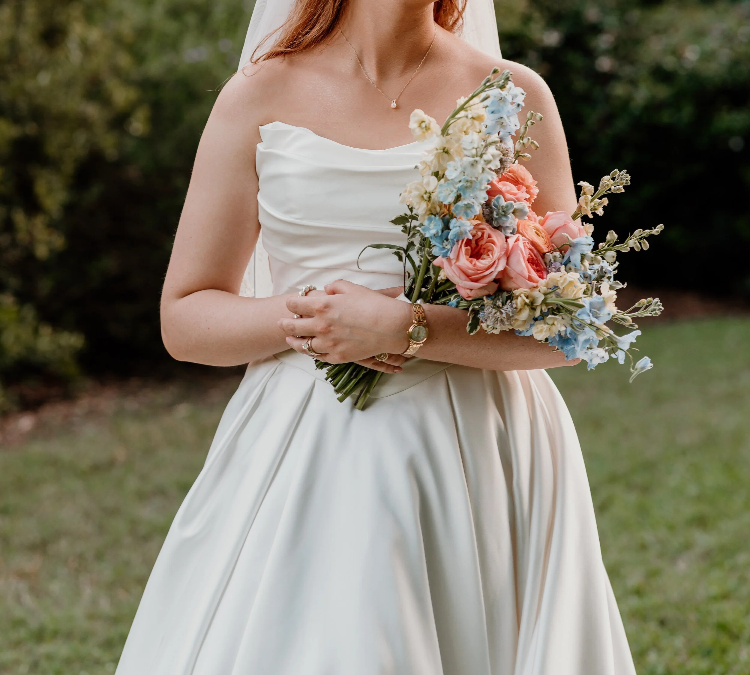 Bride in an off-the-shoulder gown holding a loose pastel bouquet of peach, pink and pale blue flowers in a garden (flower types unsure)