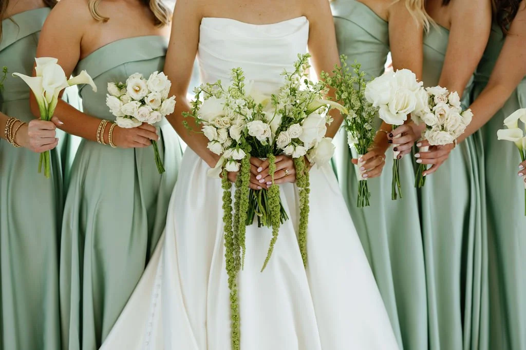 A detail of a bridal bouquet with white garden roses, lisianthus, and trailing green hanging amaranthus, and the bridesmaids holding single flower bouquets of roses, garden roses, lilies