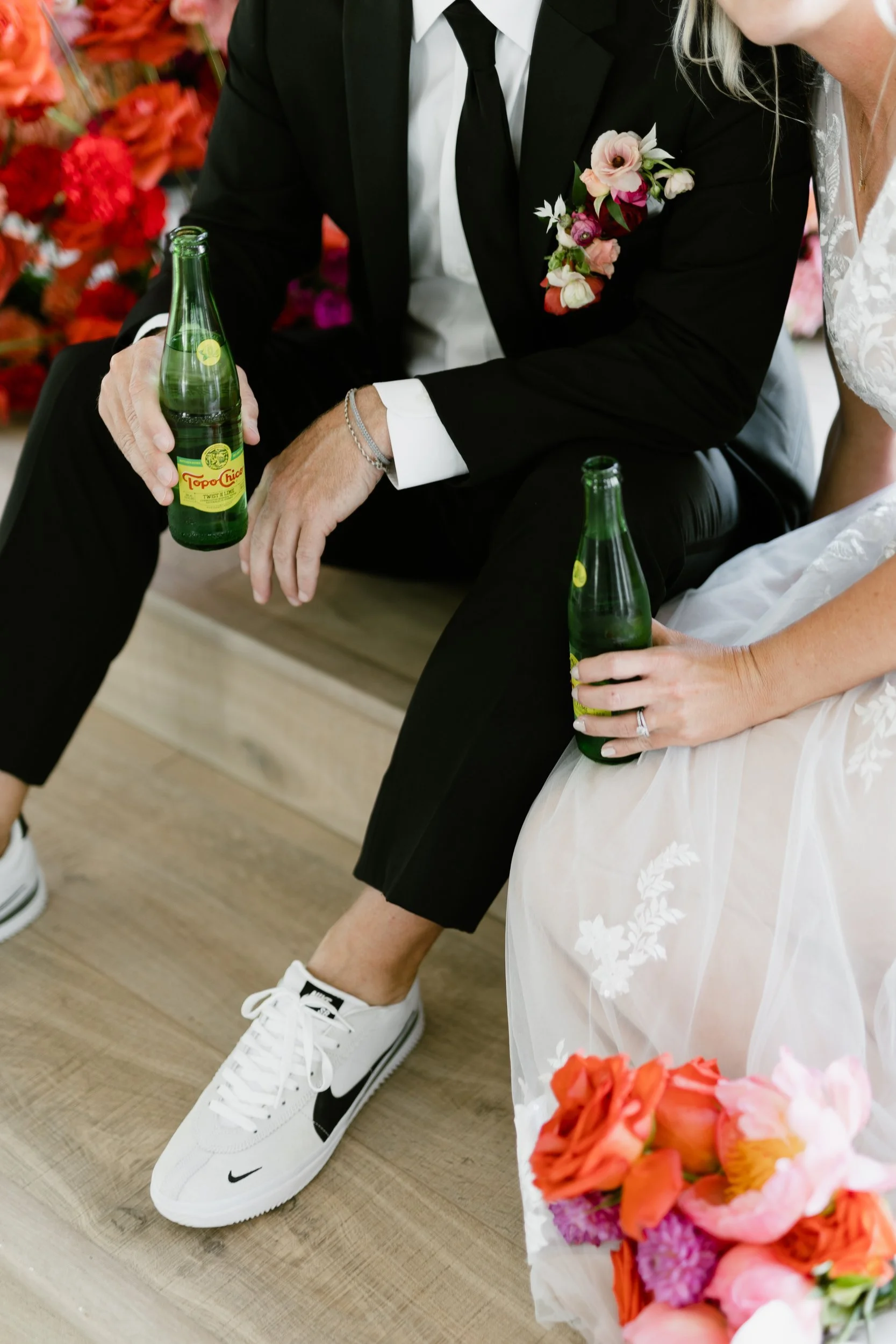 Seated couple holding drinks, groom wearing a boutonniere and a coral and pink bouquet resting at their feet