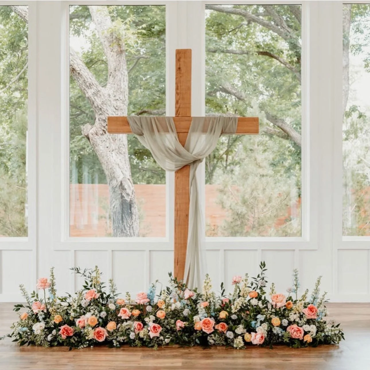 Wooden ceremony cross draped in sage fabric with a ground arrangement of peach roses and garden flowers by Haute Poppies
