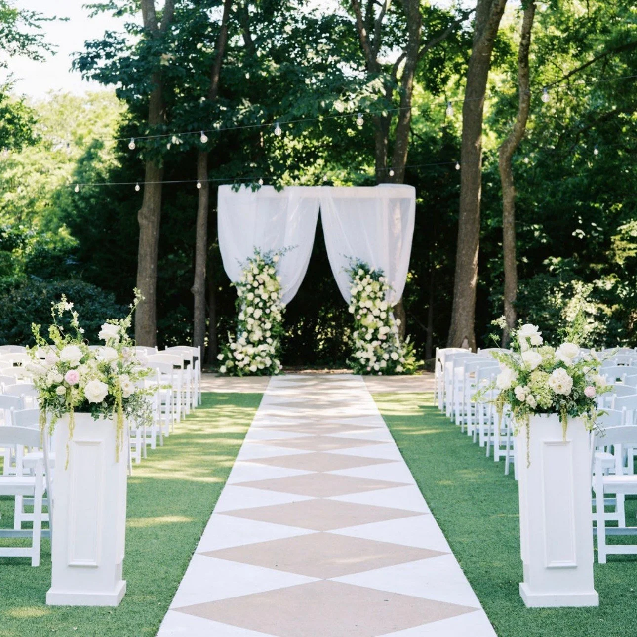 Outdoor ceremony aisle with a checkered runner, white pedestal arrangements and floral pillars framing a draped arbor under trees (flower types unsure)